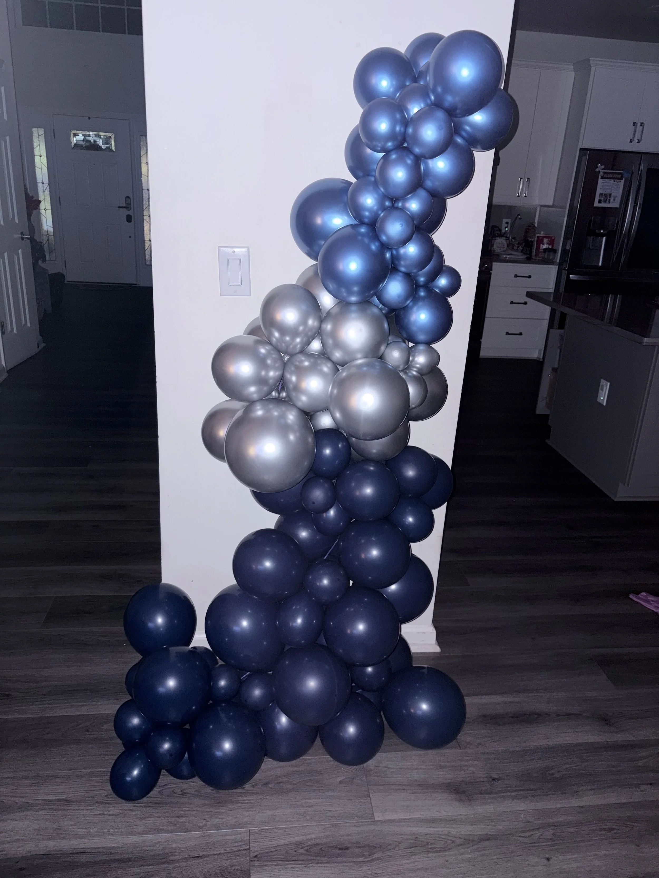 Arrangement of blue and silver balloons clustered together, placed on a wooden floor in a home interior.