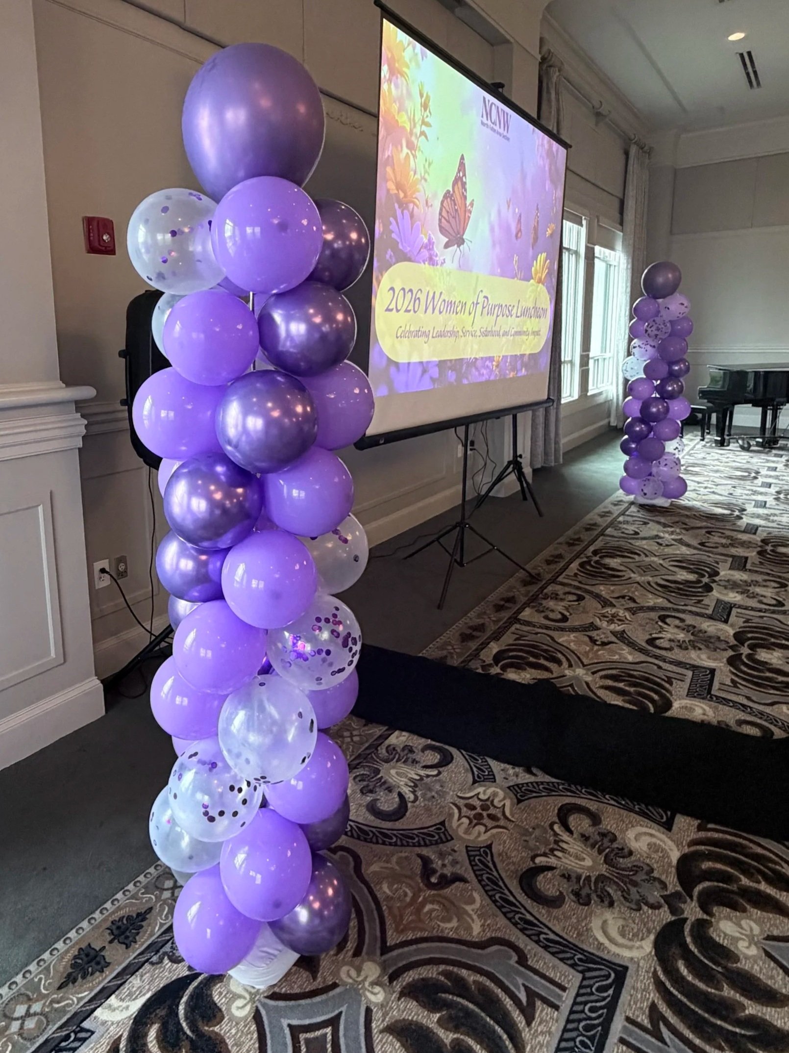 Purple balloon columns with metallic and transparent balloons, decorating a conference room with a projector screen displaying a 2026 Women of Purpose Luncheon event slide, in an elegant setting with windows and a patterned carpet.