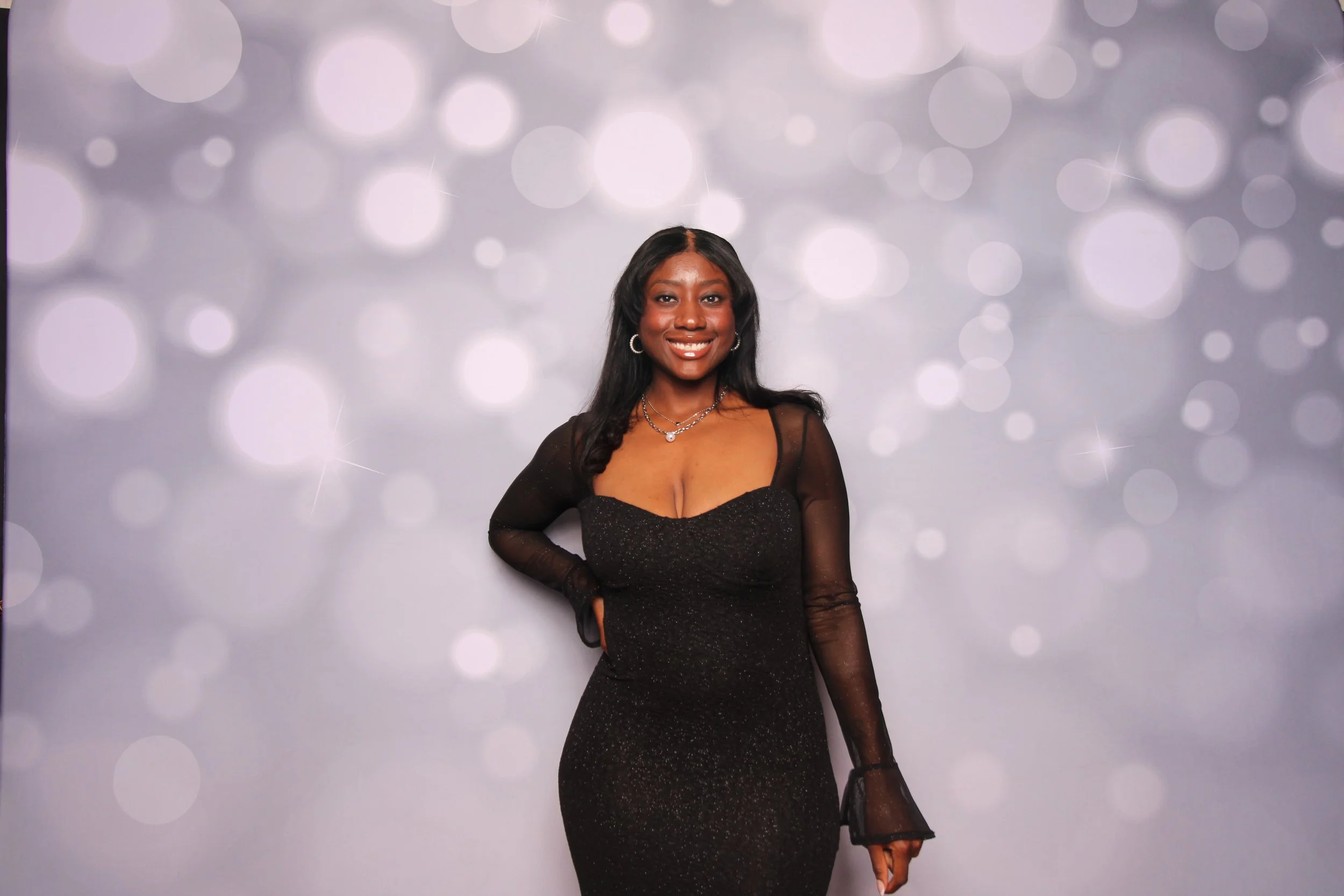 A woman wearing a black dress with sheer long sleeves, standing in front of a bokeh light background, smiling at the camera.