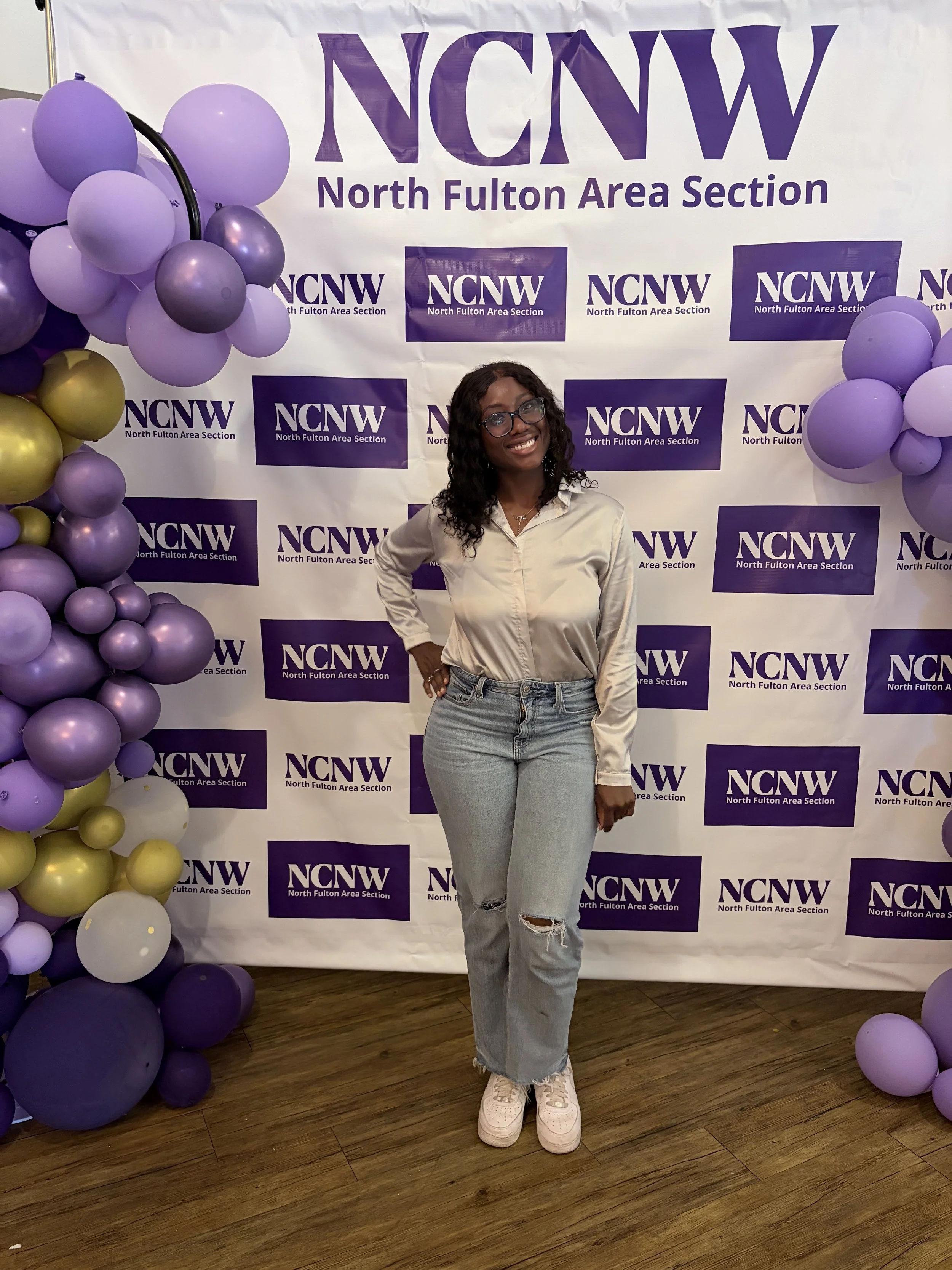 A woman standing in front of a backdrop with the NCNW logo and banner, surrounded by purple and gold balloon decorations, at an event for the North Fulton Area Section.