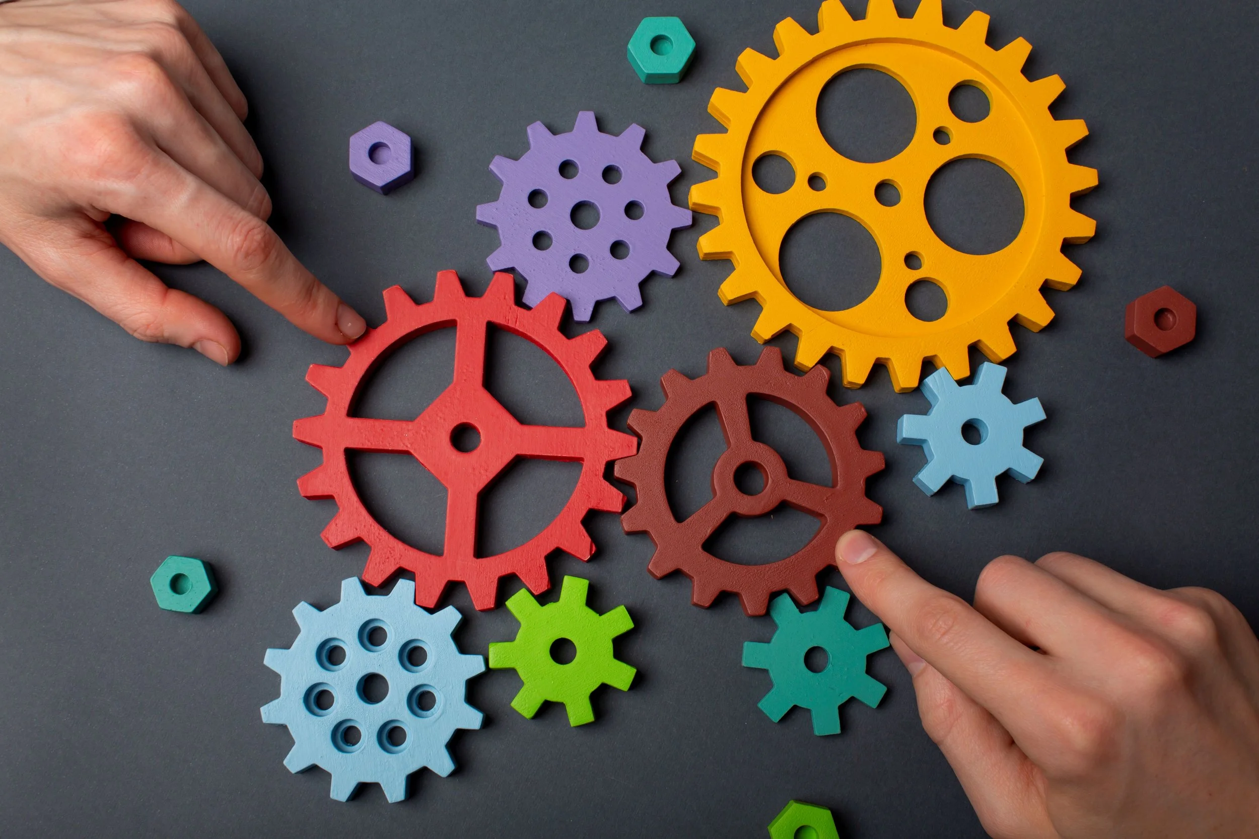Colorful interlocking gears and hands assembling them on a dark surface.