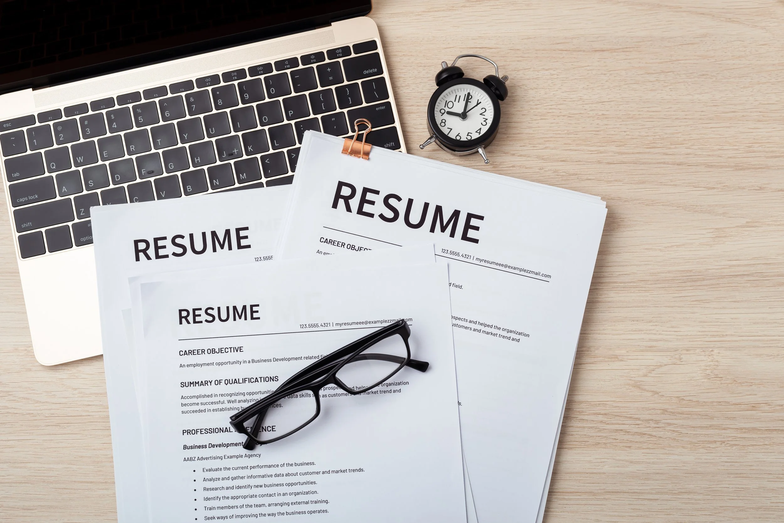 Printed resumes, a pair of glasses, a small black alarm clock, and a laptop on a light wooden desk.
