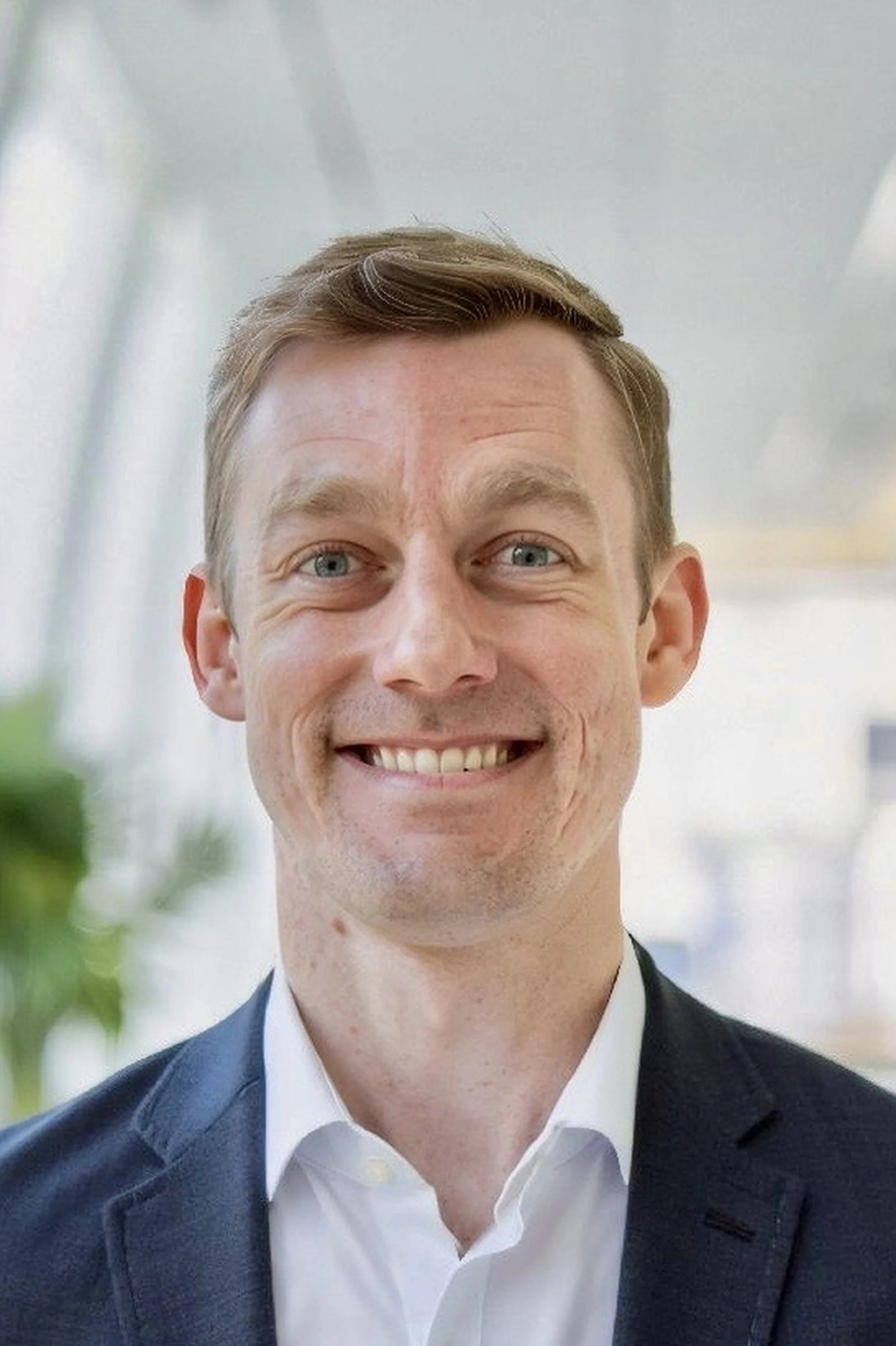 A smiling man with blue eyes and short light brown hair, wearing a light blue button-up shirt, standing outdoors with a blurred green background.