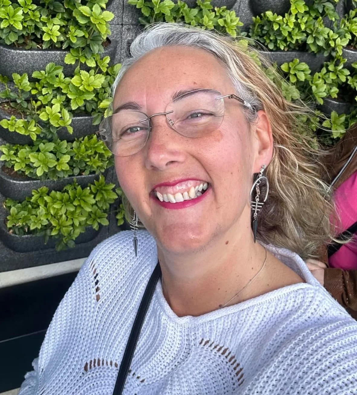 A smiling woman with glasses and earrings taking a selfie outdoors with green plants behind her.
