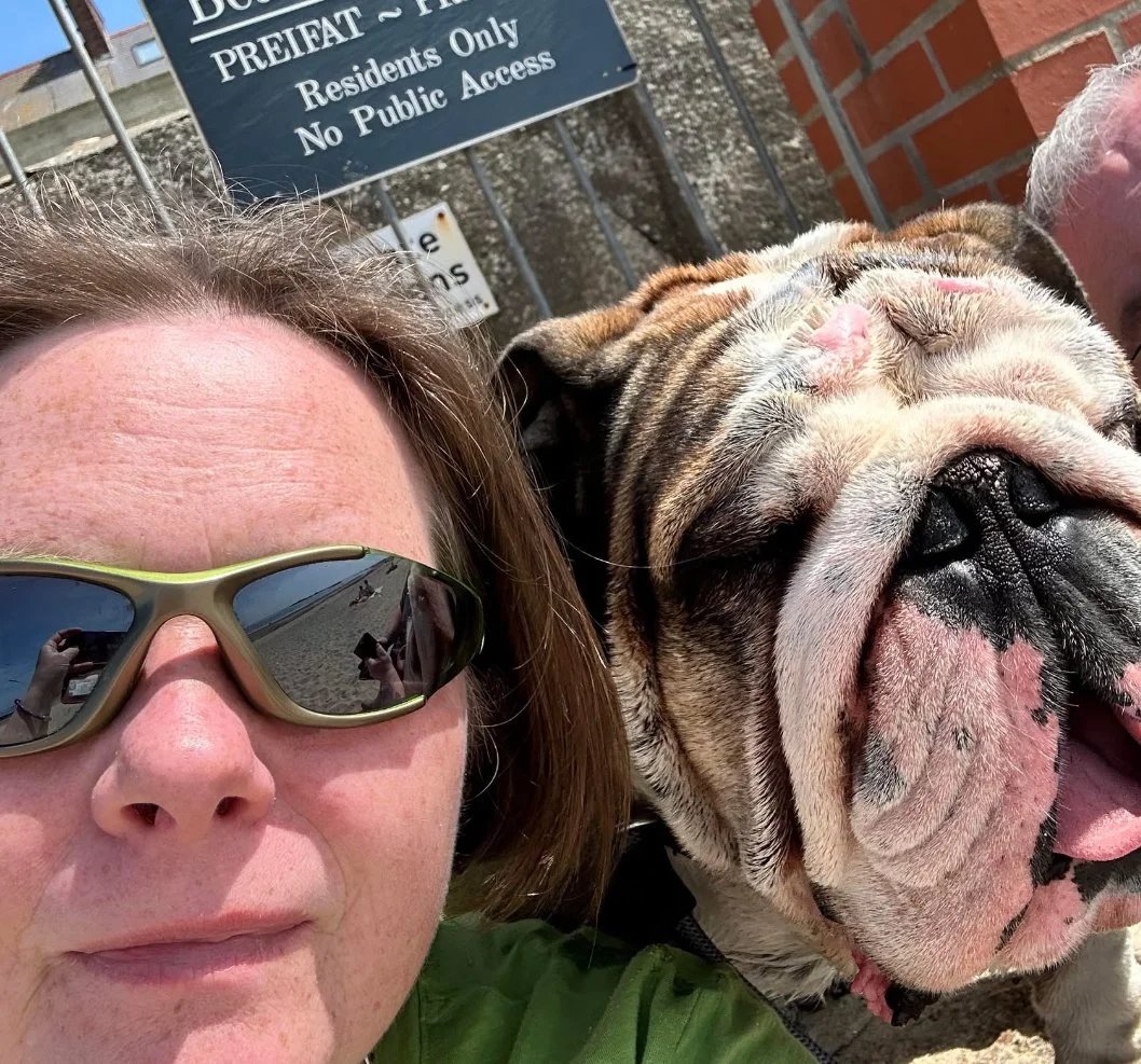 A woman with short brown hair wearing sunglasses taking a selfie with a large bulldog dog. The background includes a metal fence, a brick wall, and a sign that reads 'Residents Only No Public Access'.