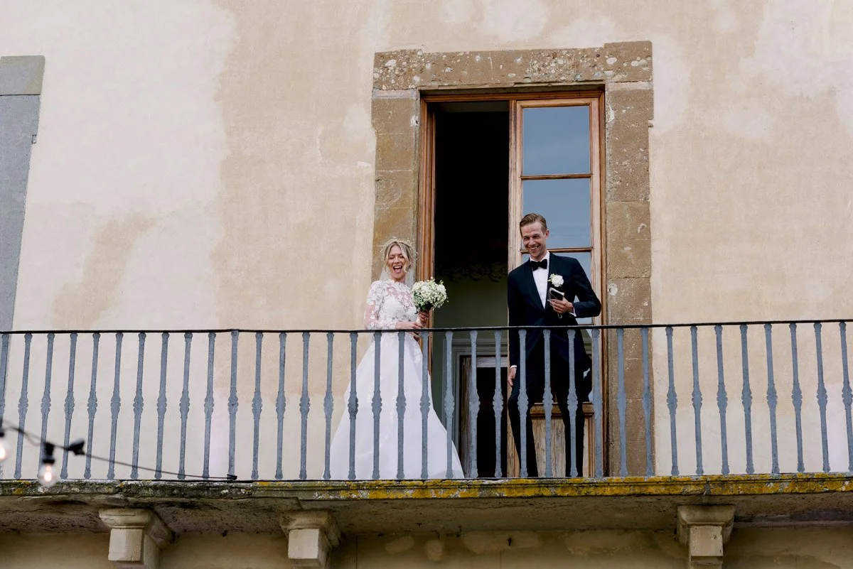Bruidspaar op het balkon van een Toscaans kasteel, lachend en genietend van het uitzicht – een ontspannen moment tijdens een perfect georganiseerde dag met ceremoniemeester, een prachtig voorbeeld van trouwen in Toscane.