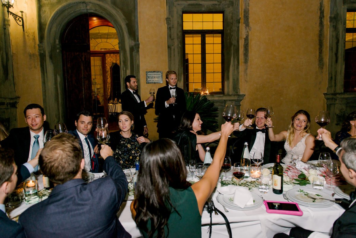 Bruidspaar en gasten proosten met een glas wijn tijdens het bruiloftsdiner bij Castello di Montegufoni – een vrolijk en zorgeloos moment dankzij de ceremoniemeester en perfecte planning in Toscane.