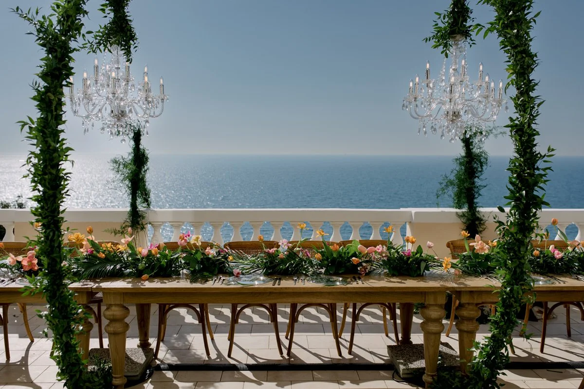 Lange dinertafel met bloemen en kroonluchters aan de Amalfikust, met uitzicht op zee, als voorbeeld van hoe sfeer en logistiek bij trouwen in Italië sterk per regio verschillen.
