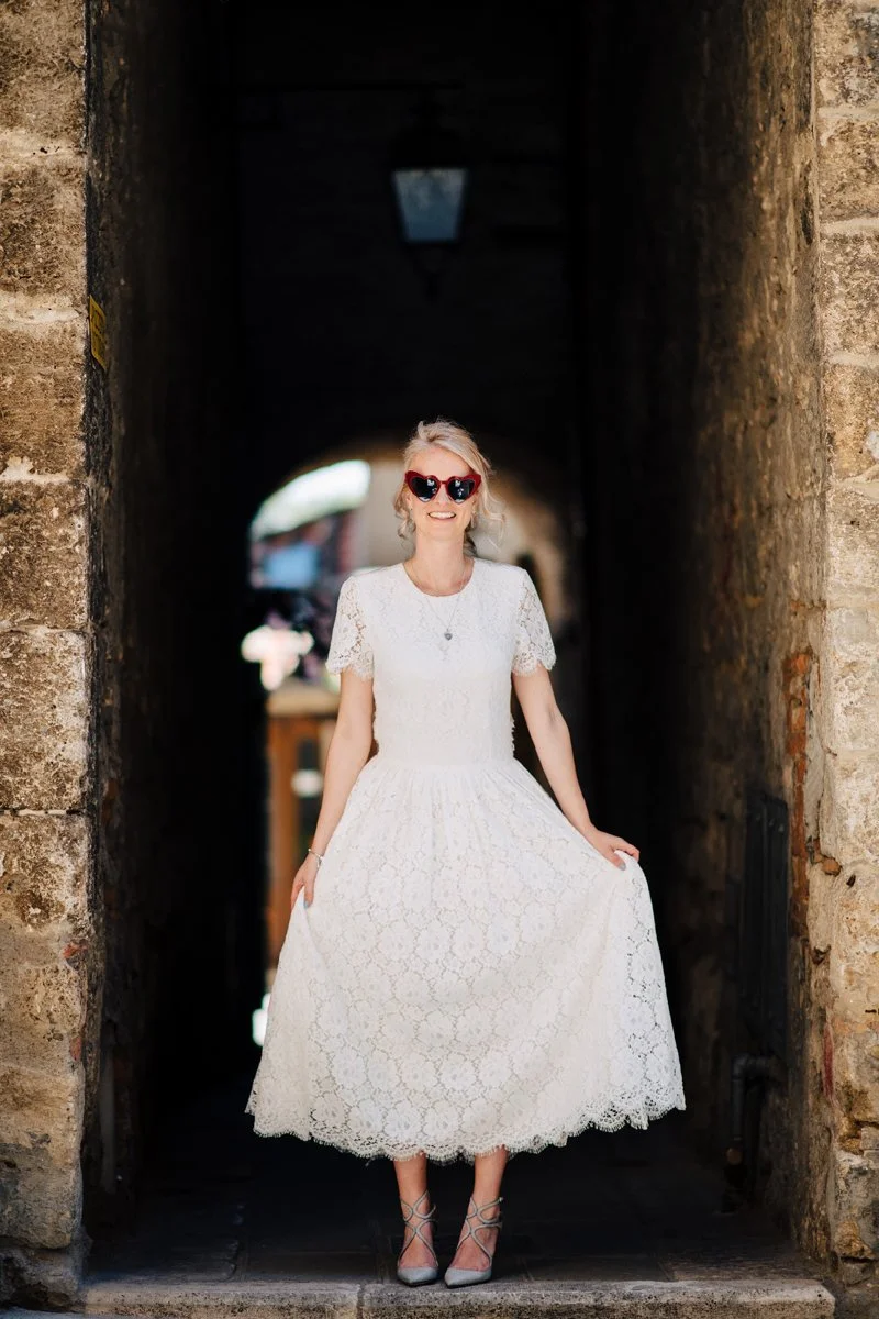 Bruid met rode zonnebril en retro jurk lacht in een smal straatje van een Toscaans stadje – een speels en stijlvol elopement moment, vastgelegd door de trouwfotograaf van Toscane.