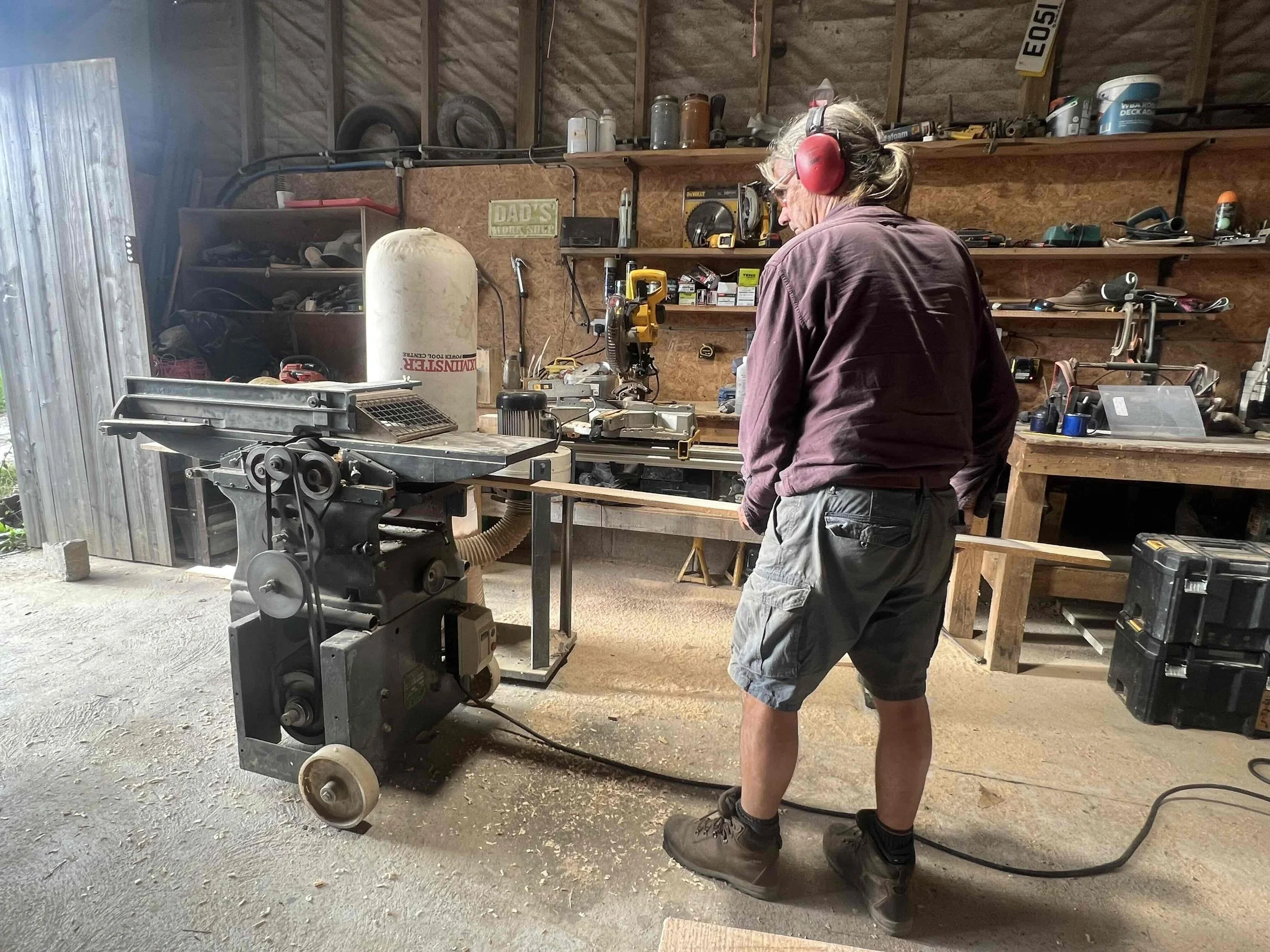 Older man working in a woodworking shop using a band saw, wearing protective ear muffs and glasses.