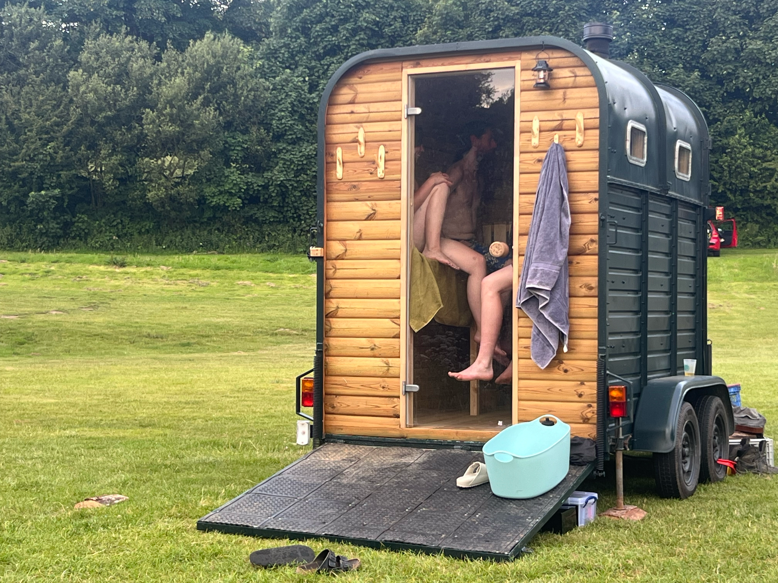 Two people sitting inside a small wooden sauna trailer outdoors, with towels hanging on the exterior, and various bath items around, including a blue basin and slippers on a ramp leading inside.