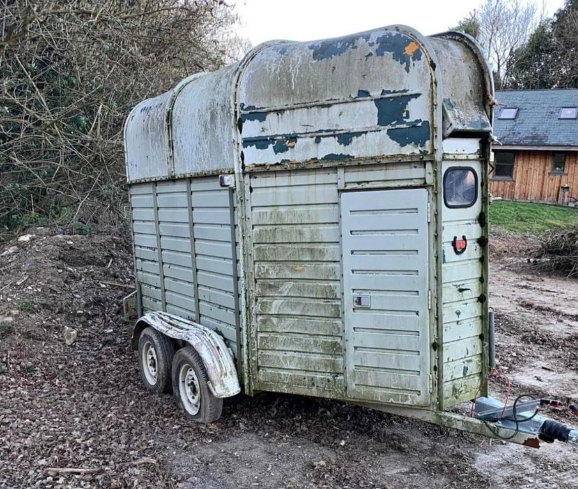 Old, rusted, and weathered small horse box with two wheels, parked on dirt outdoors near a shrub and a wooden house.