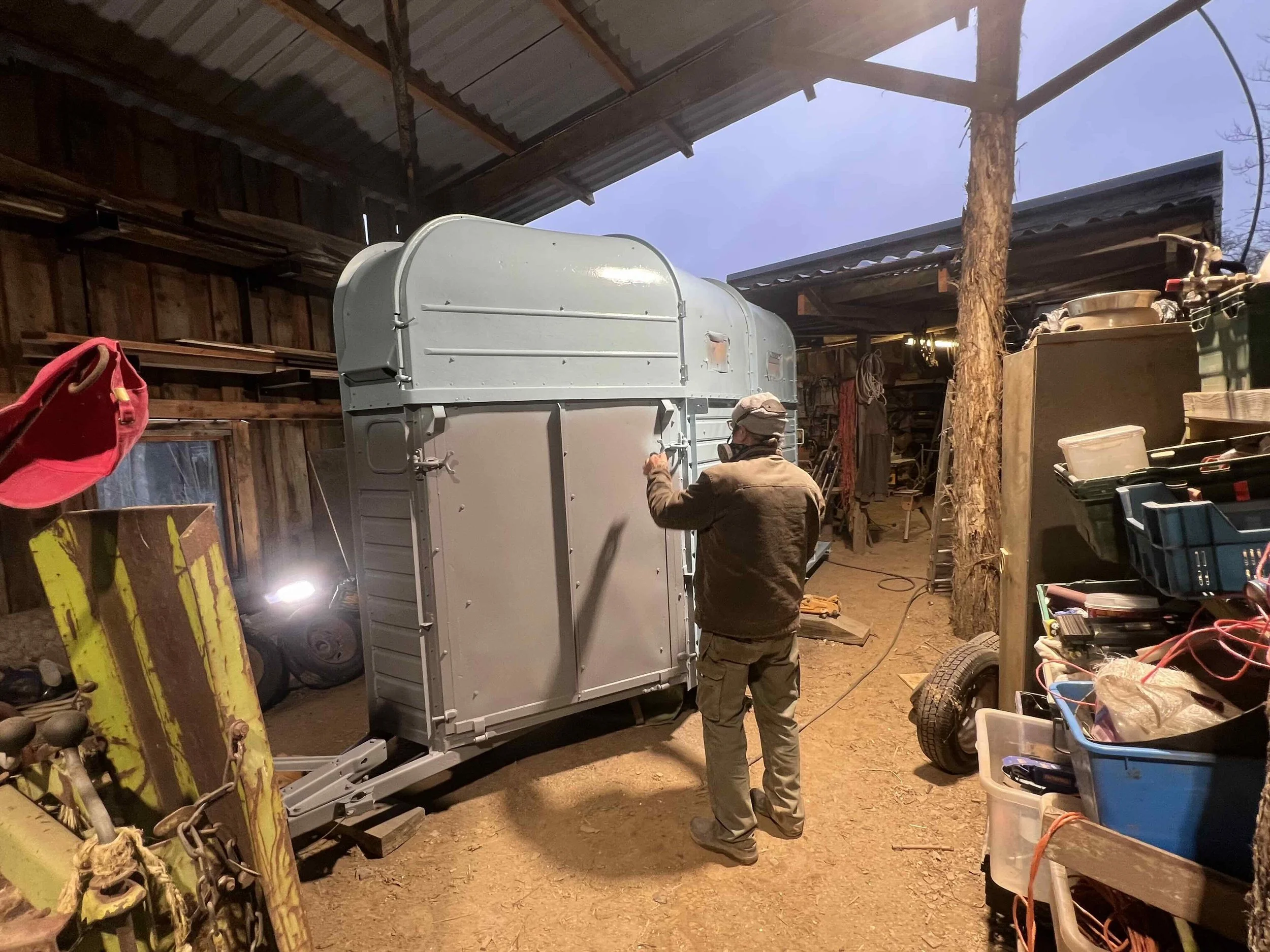 A person standing inside a barn or shed, working on a new metallic trailer or container. The barn has wooden walls and a rustic interior with various tools and equipment around.