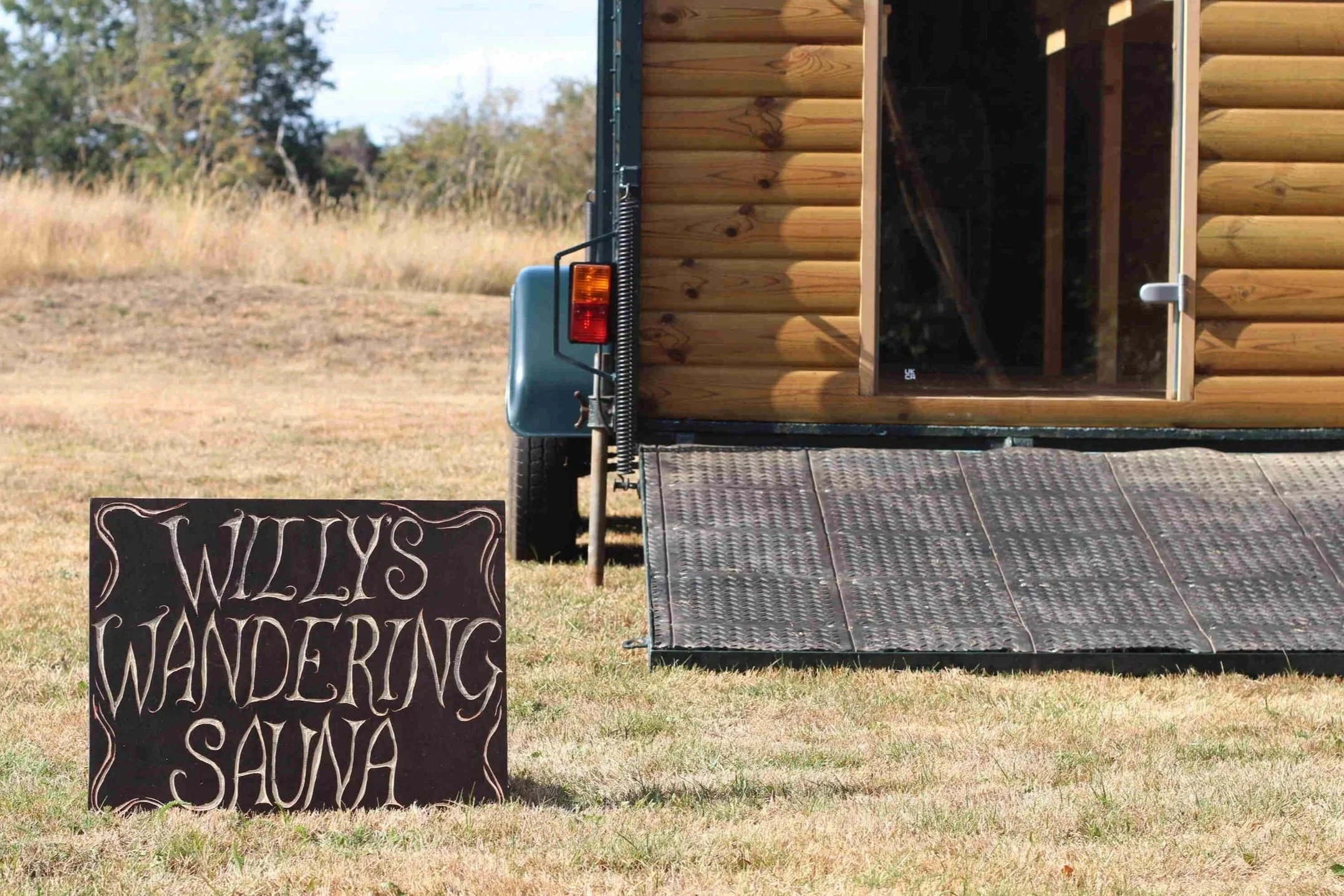 A small wooden sauna with an open door, positioned on a grassy field. In front of the sauna, there is a chalkboard sign that reads 'Willy's Wandering Sauna' in decorative writing. Taken before a wedding in Devon.