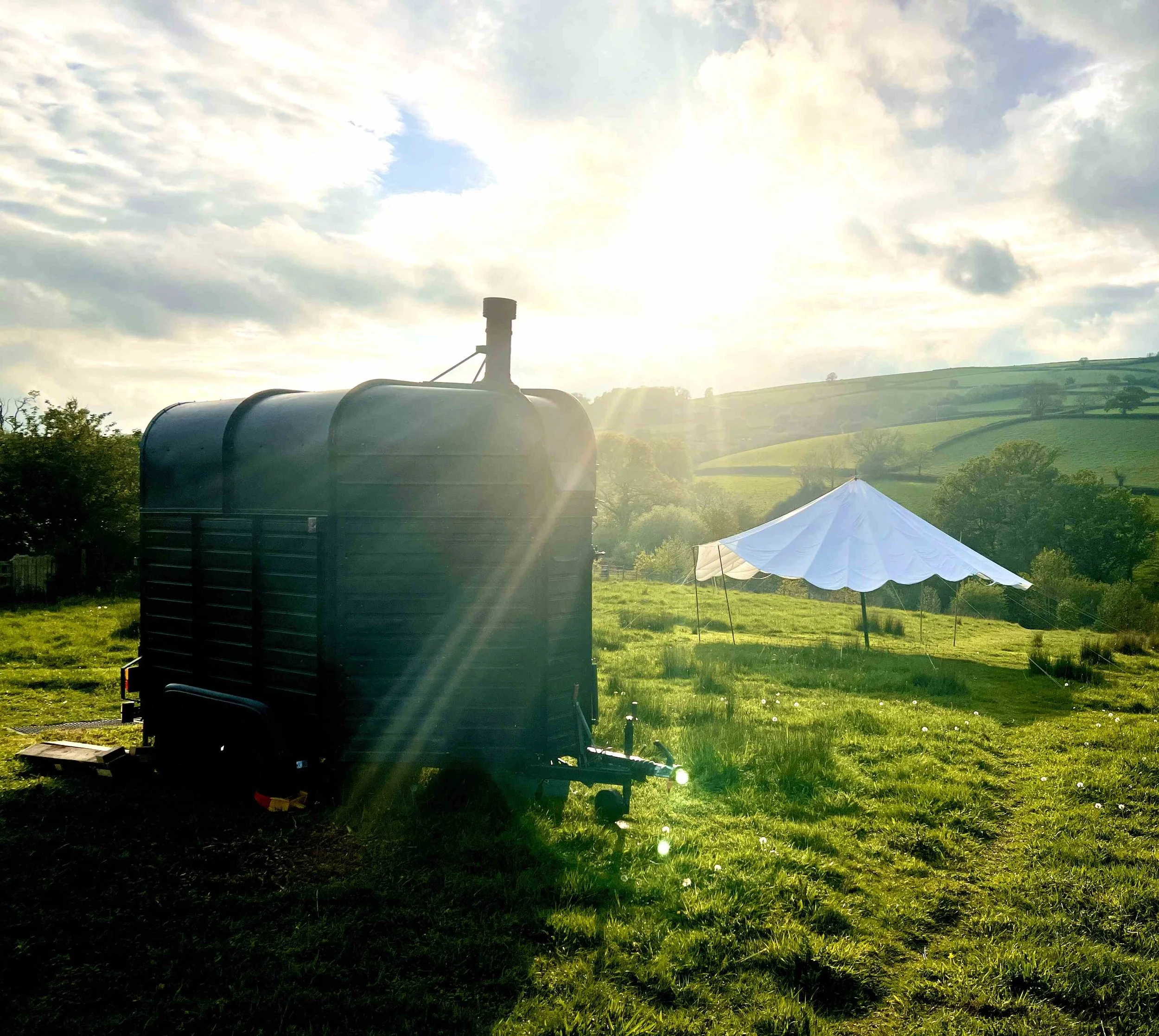 A large black water tank sits in a grassy field with a white canopy tent nearby. Hills and scattered trees are in the background under a partly cloudy sky with the sun shining.