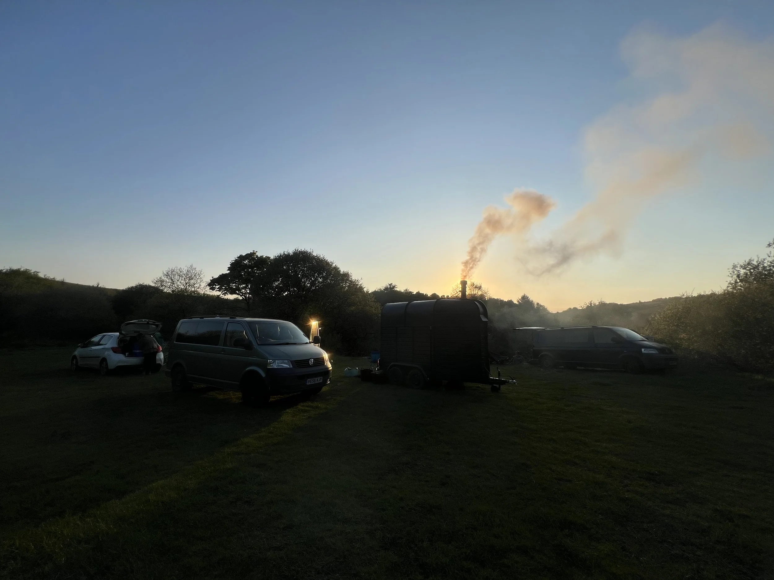 A sunset scene with cars parked on grassy land, a small mobile sauna by the river on Dartmoor, and smoke rising from a chimney in the distance.