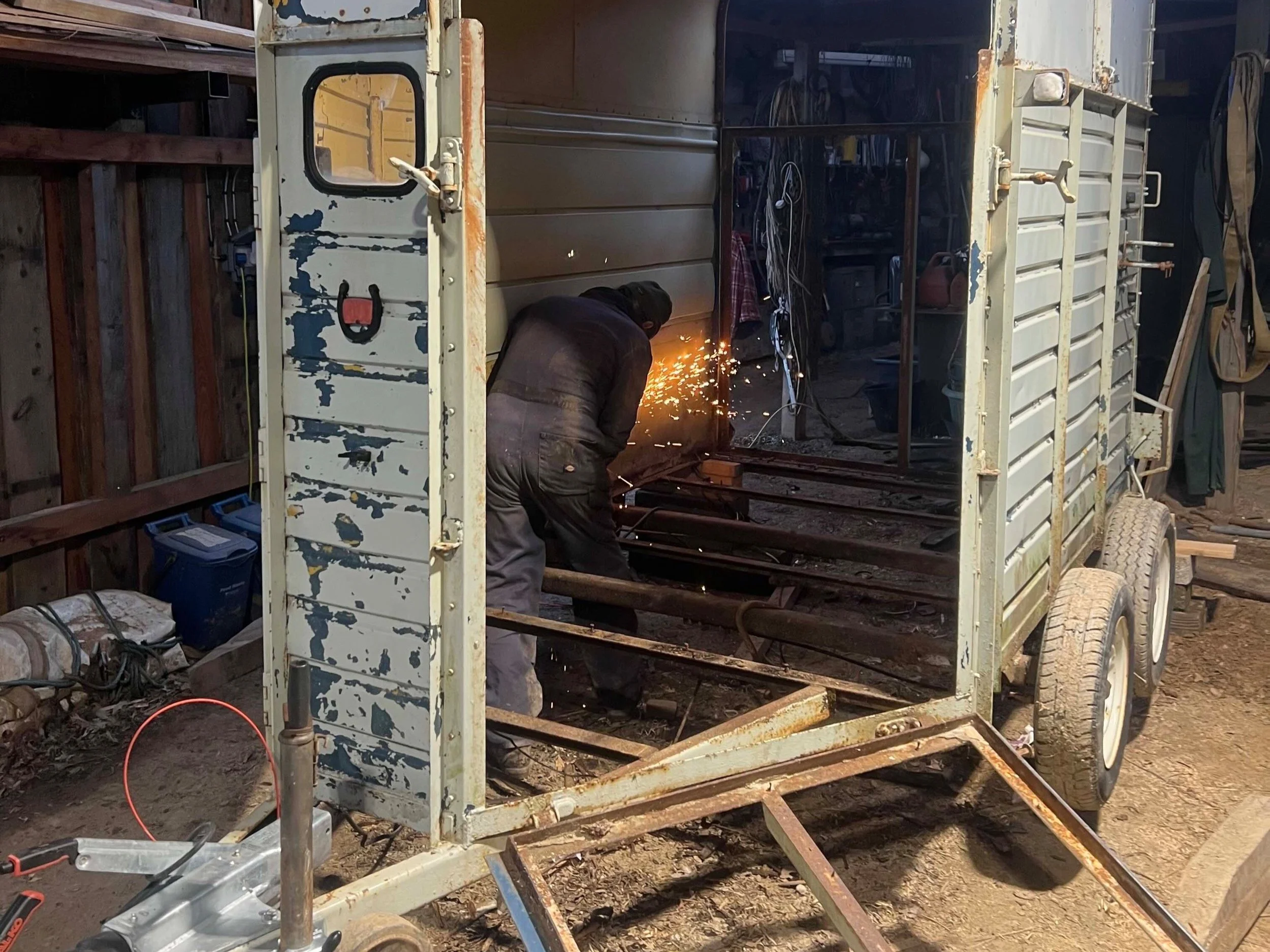 A person welding metal inside a small, partially assembled trailer with rusted exterior in a workshop.