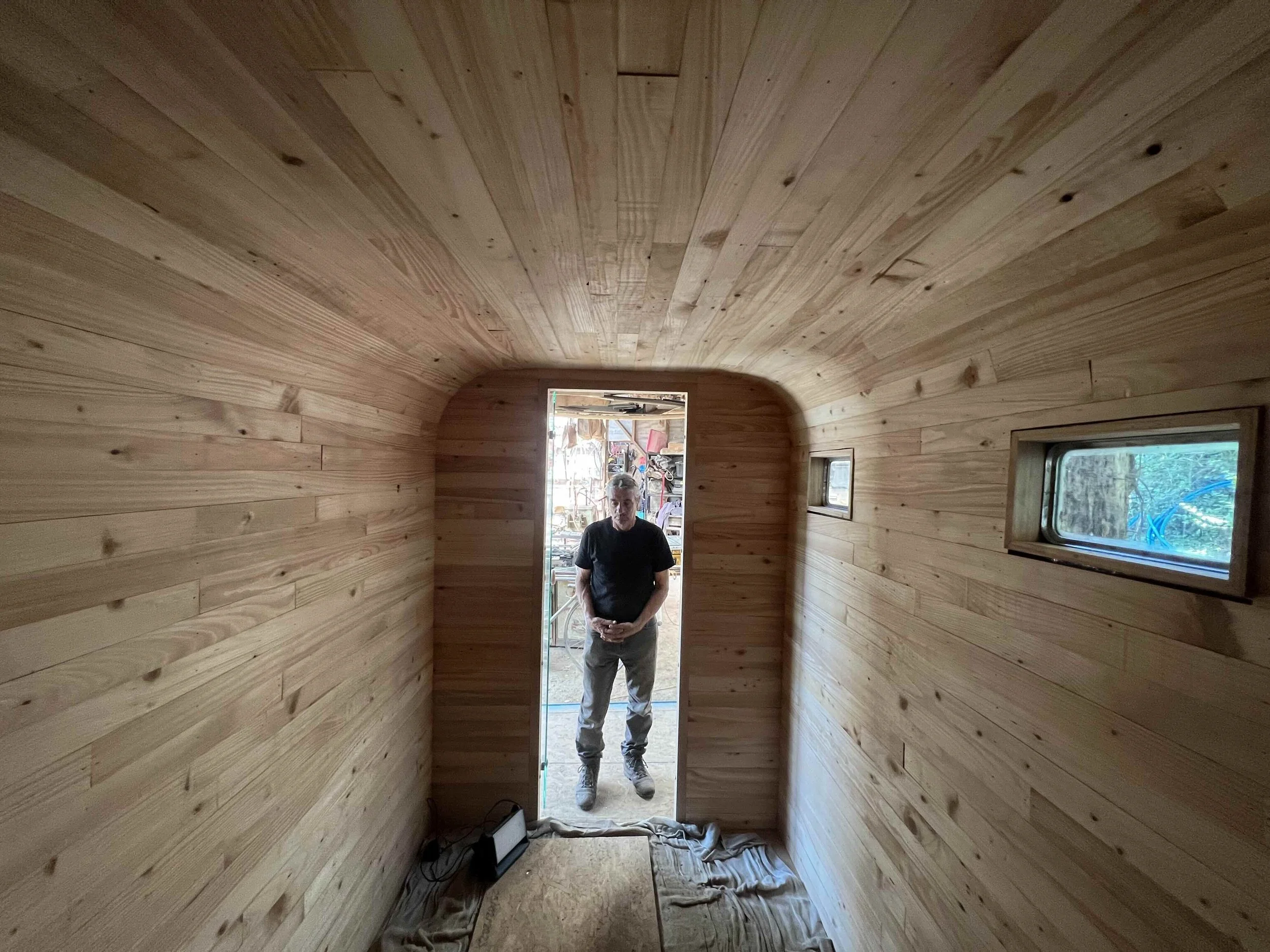 A man standing in a narrow, wooden, tunnel-like room with small windows on the side. The walls and ceiling are made of light-colored wooden planks. The doorway shows an outdoor workshop area with tools and materials.