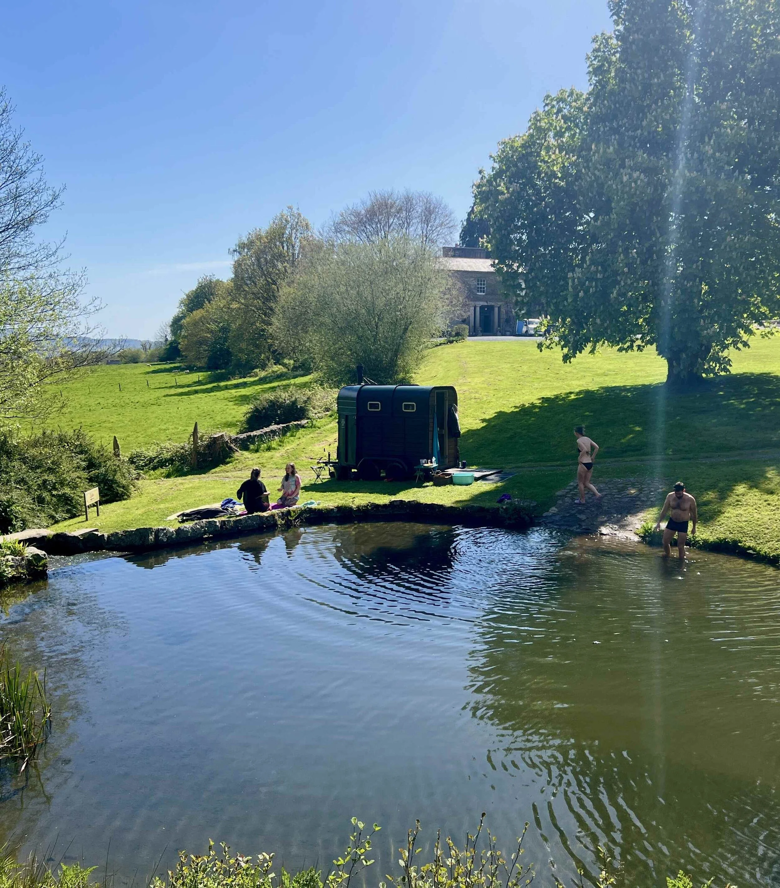 A peaceful outdoor scene with a pond in the foreground reflecting the sky and trees. People are enjoying leisure activities on the grass, and a country house is visible in the background amidst lush trees under a clear blue sky.