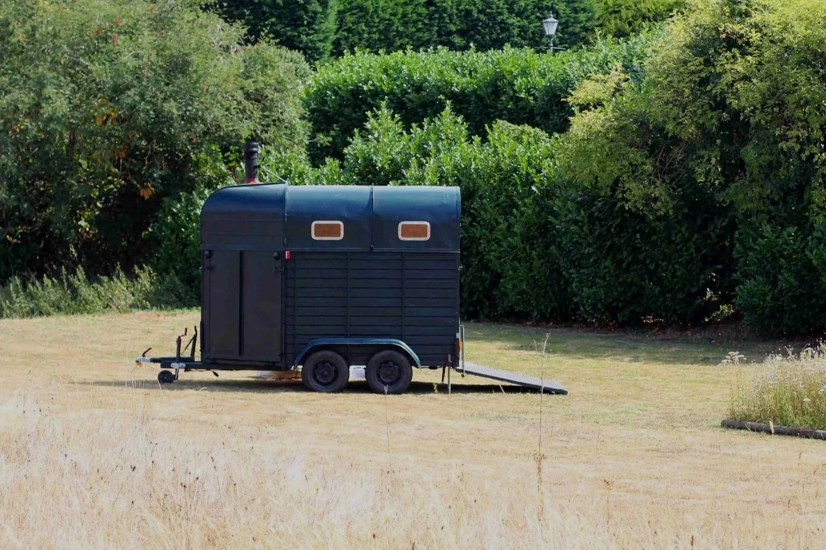 A small black mobile sauna with a ramp parked on a grassy area surrounded by trees.