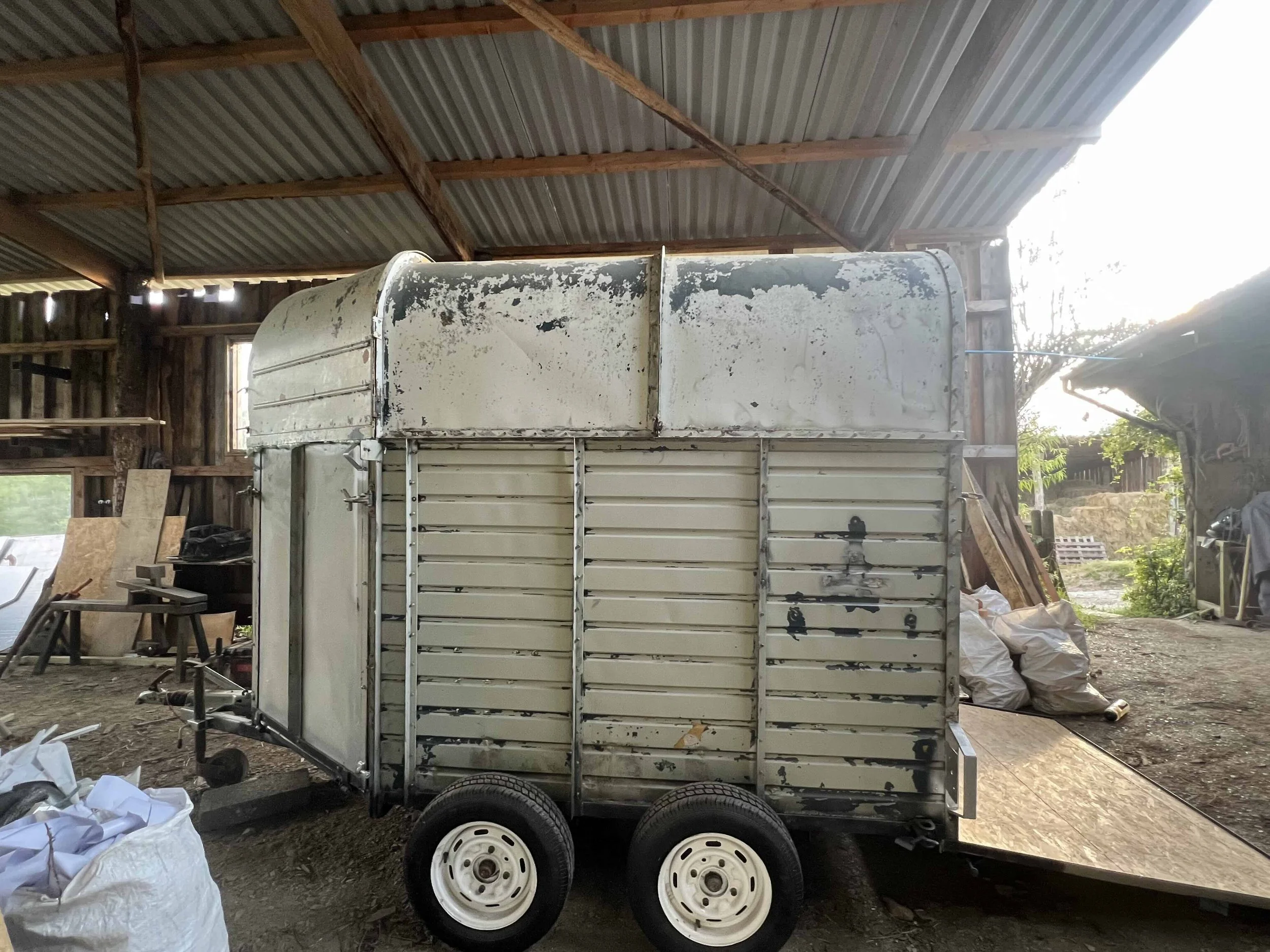 An old, weathered, white metal trailer with two wheels, parked inside a wooden barn with a corrugated metal roof. The trailer shows signs of rust and paint peeling, and is connected to a ramp on its right side. Various items and bags are scattered ar