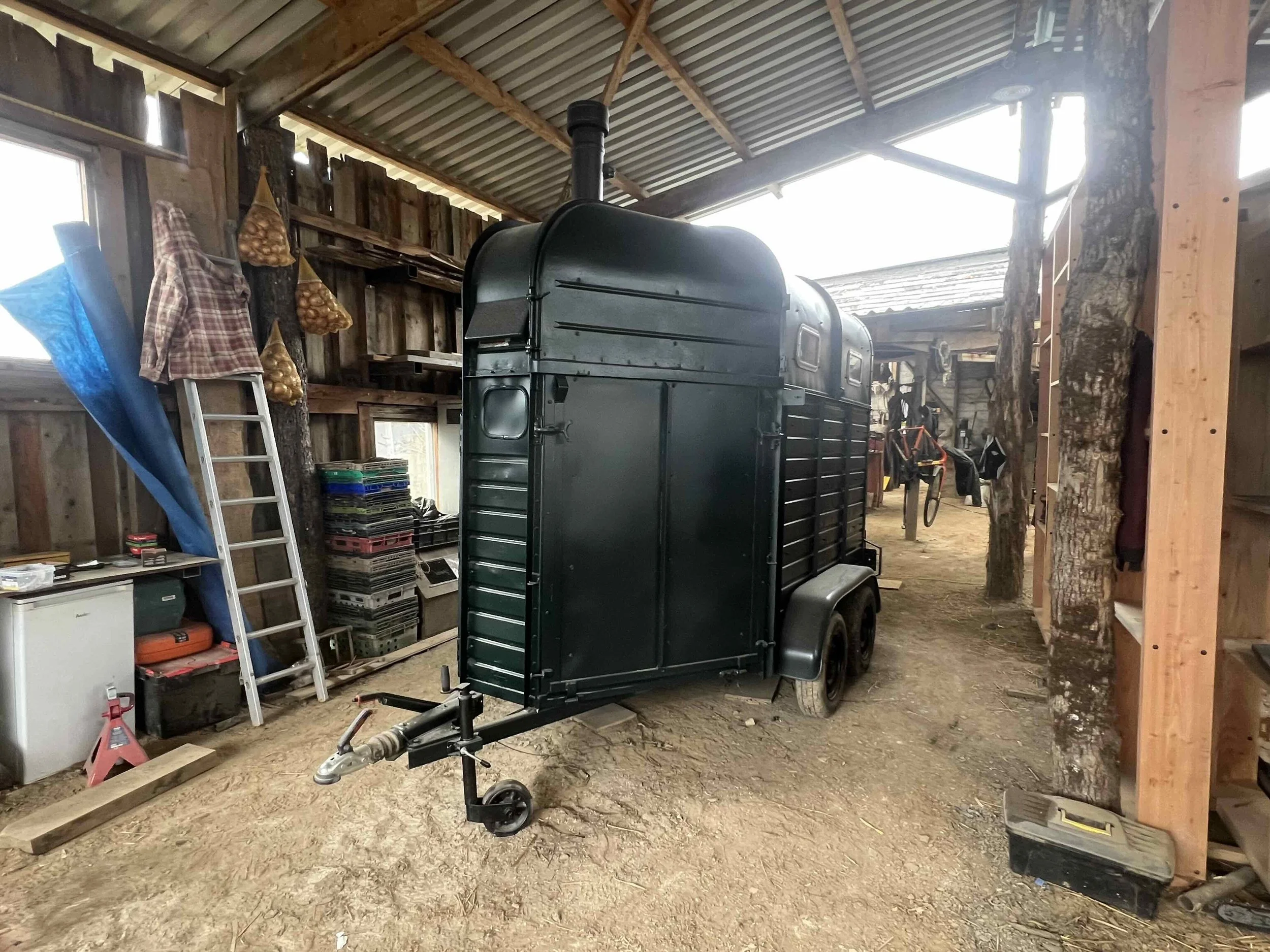 A black horse trailer inside a rustic barn with a dirt floor, wooden walls, and a metal roof. It has a hitch in the front and is surrounded by various farm tools and equipment.
