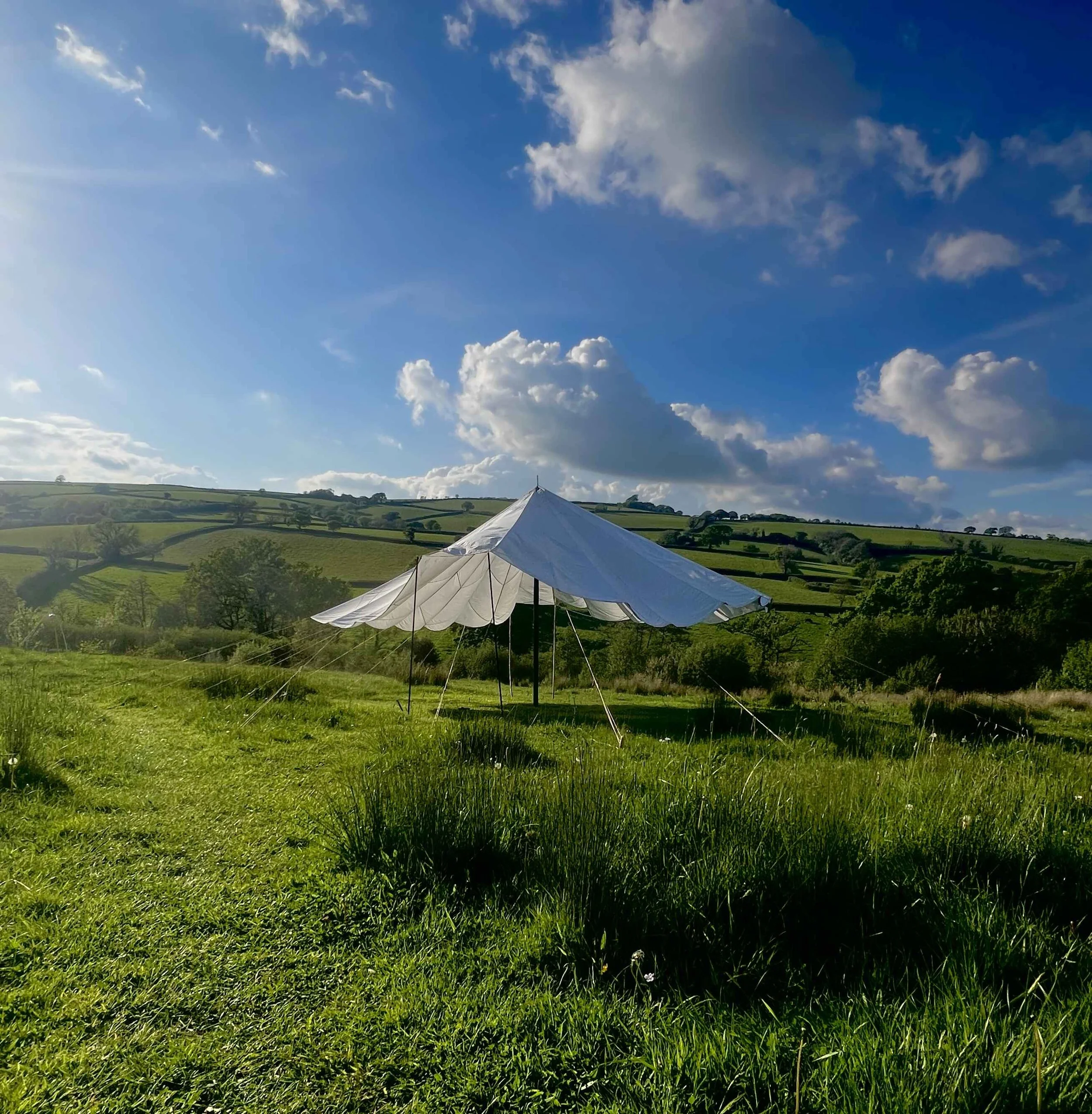 A white tent set up on a green grassy field with rolling hills and trees in the background under a bright blue sky with scattered clouds.