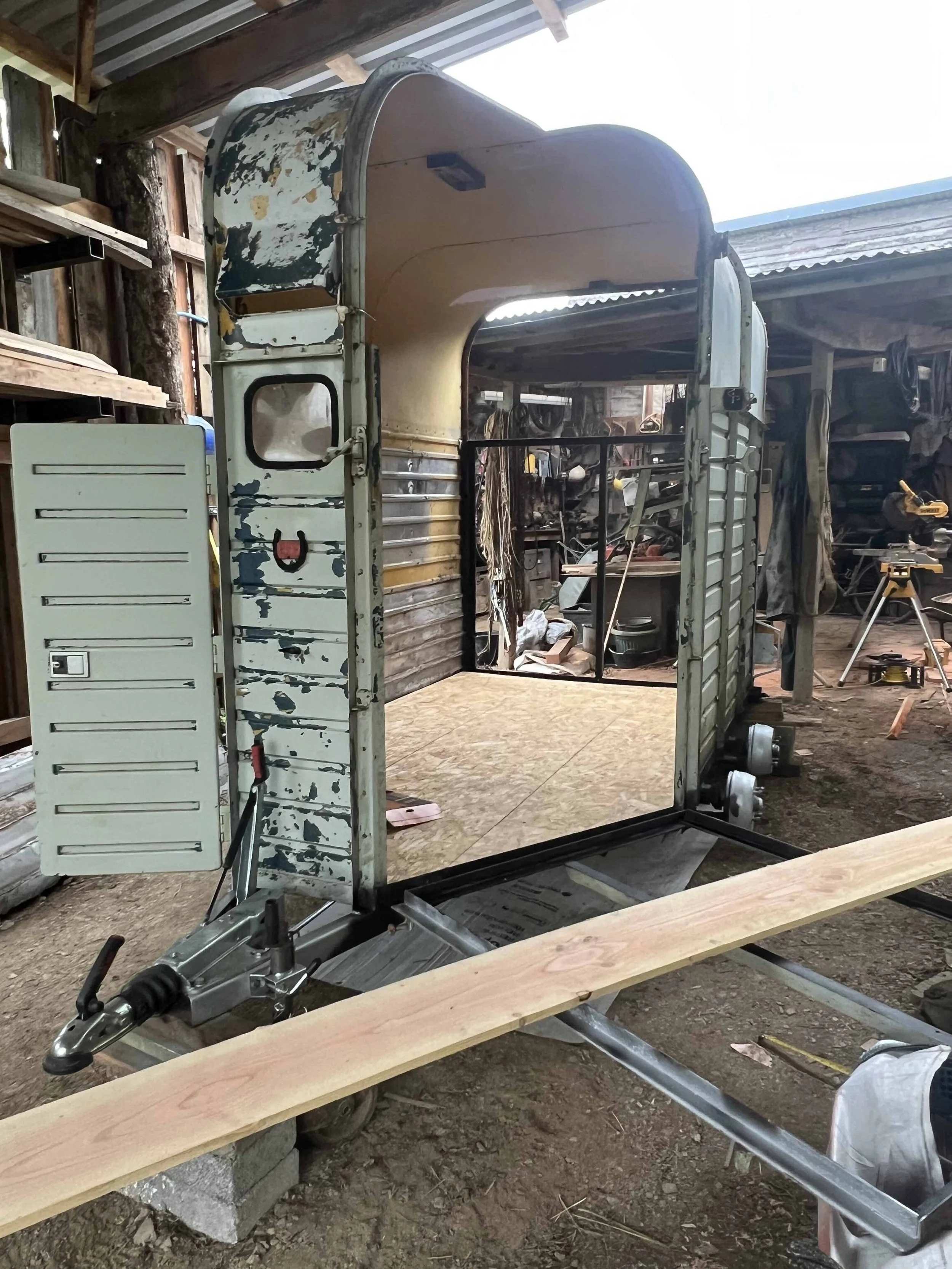 The image shows an old, partially disassembled horse boxin a workshop or garage. It has peeling paint and is mounted on a metal frame, with a piece of wood placed at the front. The surrounding environment includes tools and workbenches.