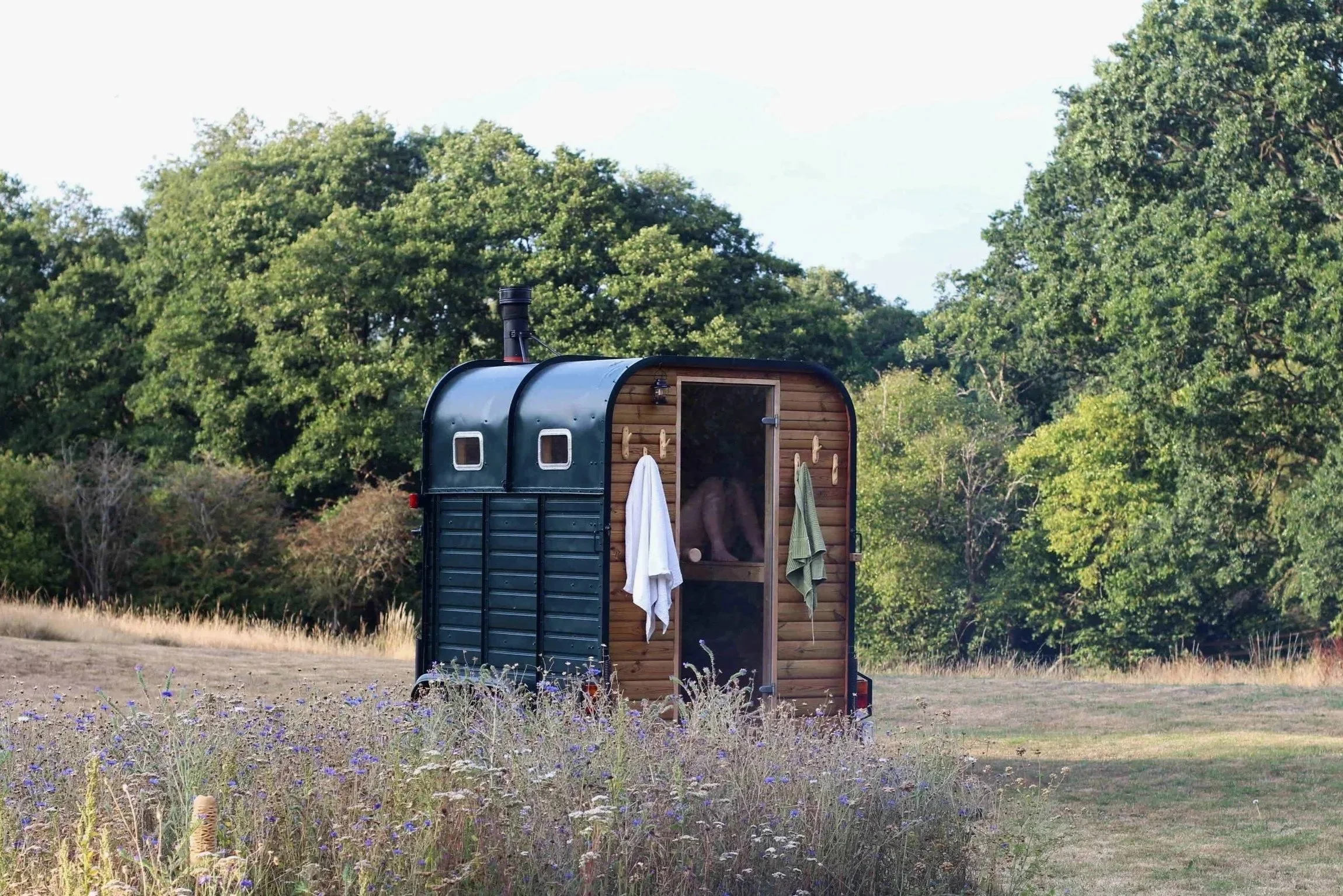 A small black and wood sauna in a grassy field with wildflowers, surrounded by trees, under a partly cloudy sky. Looks like the Westcountry or somewhere in South West England