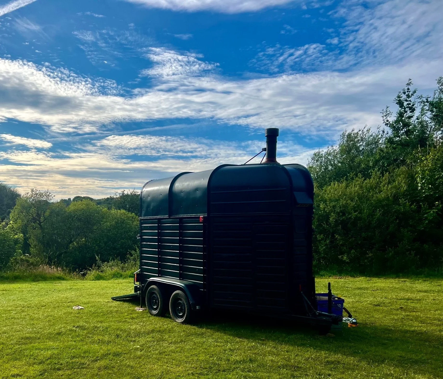 A black mobile smoker trailer on a grassy field with trees and a partly cloudy sky in the background. The picture is from the banks of the River Dart in England.
