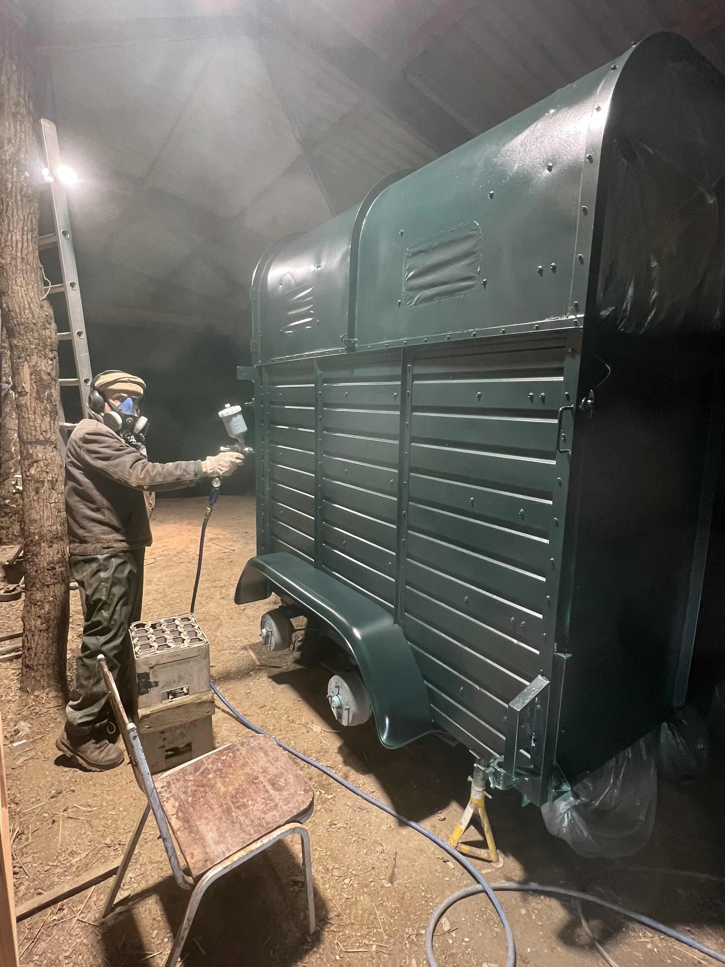 A person wearing a protective mask and gloves is spray painting a large, dark green metal trailer inside a workshop with a spray gun connected to a hose.