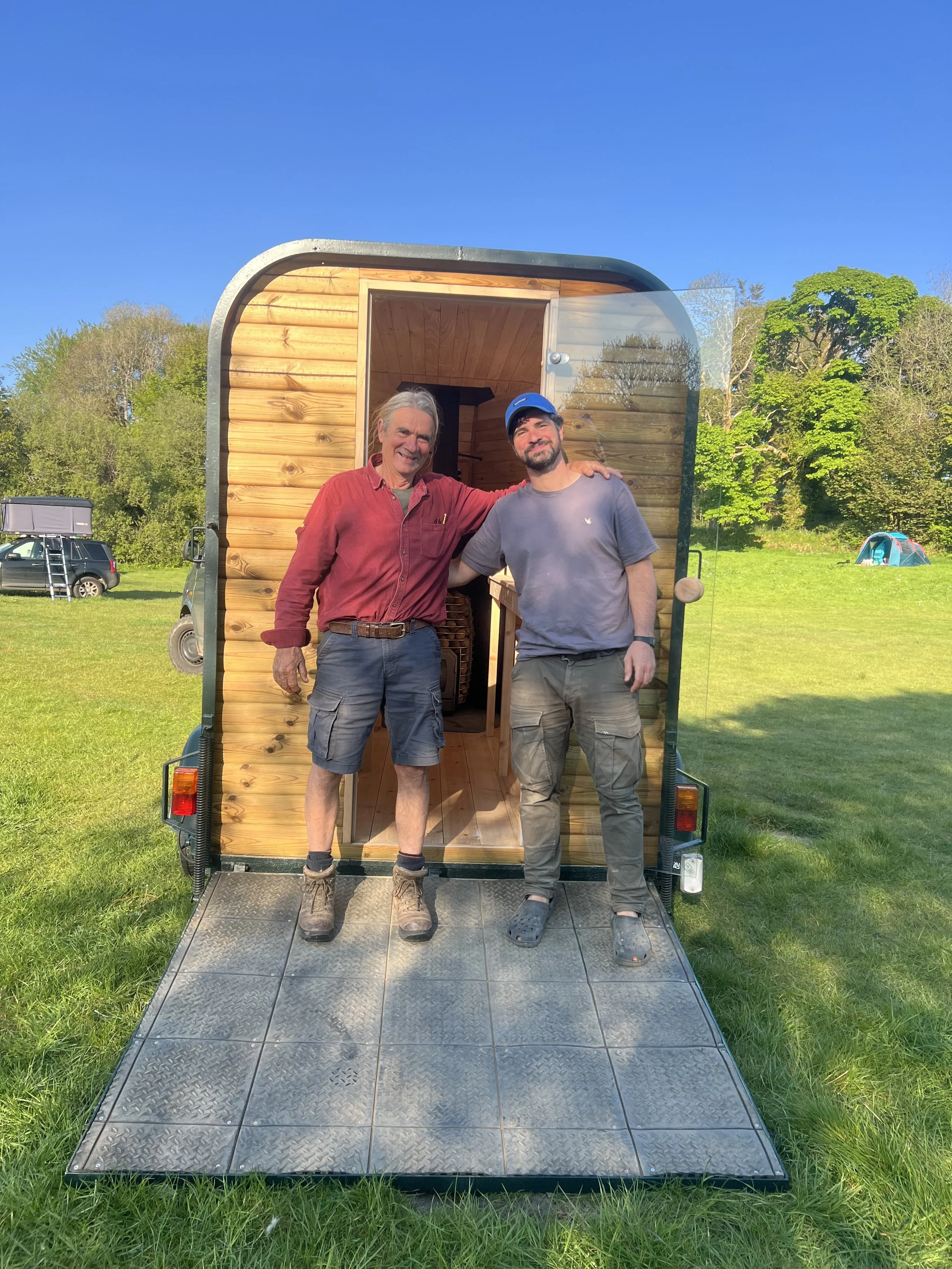 Two men standing inside a small wooden trailer with the door open, outdoors on a grassy field with trees and tents in the background.