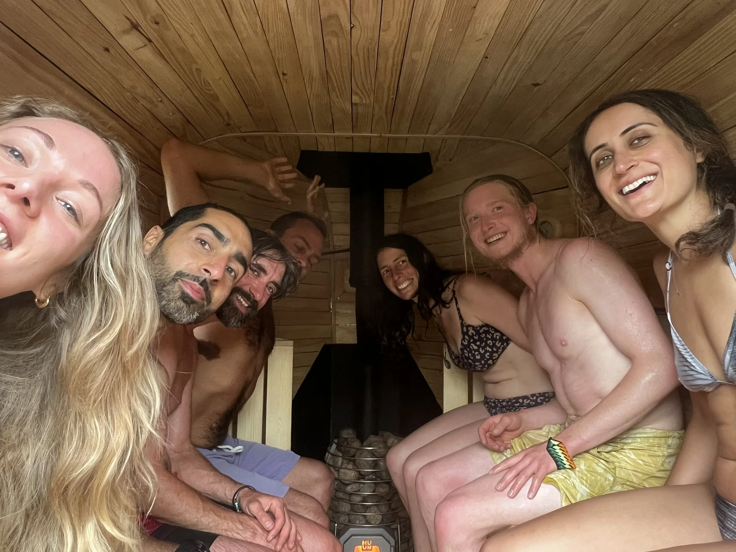 Group of seven people smiling and relaxing in a wooden sauna, sitting close together in swimwear, with a stove and rocks at the center. Taken at a birthday party in Cornwall.