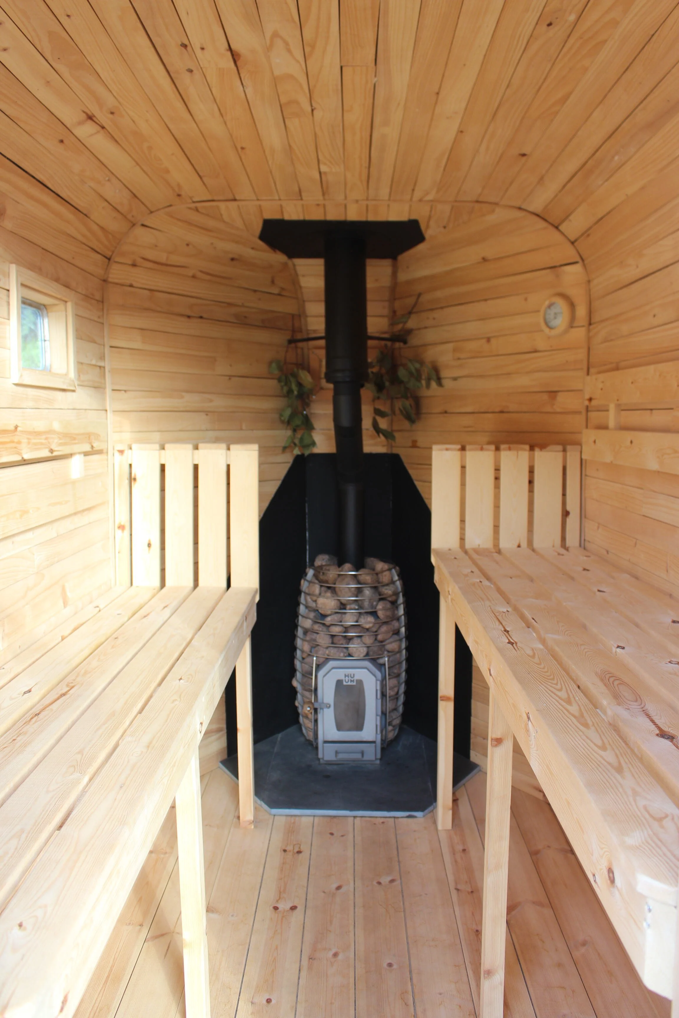 Interior of a small wooden sauna with wooden benches and a stove in the center surrounded by stones.