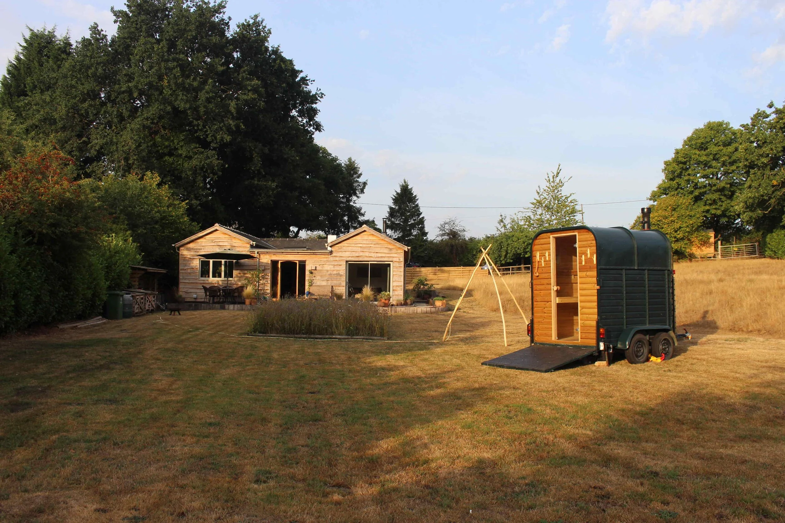 A backyard with a wooden shed, a black trailer, and a swing set in a grassy yard surrounded by trees and a fence.