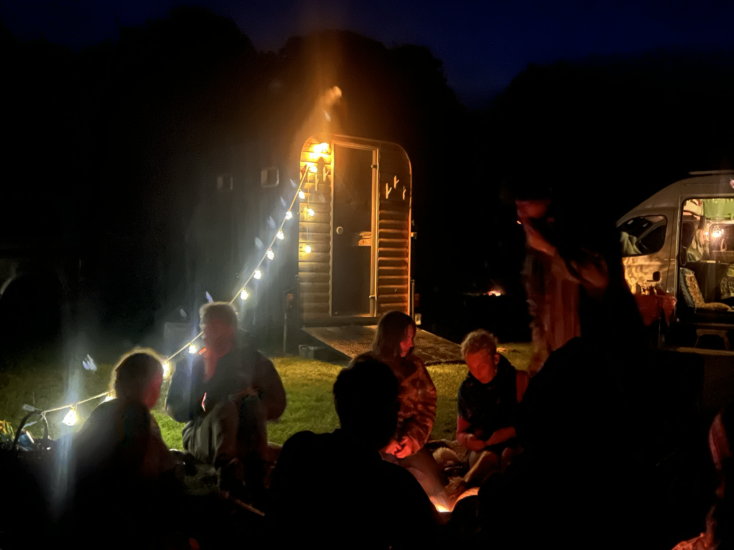 People gathered around a campfire at night, with a trailer or camper in the background, illuminated by string lights.