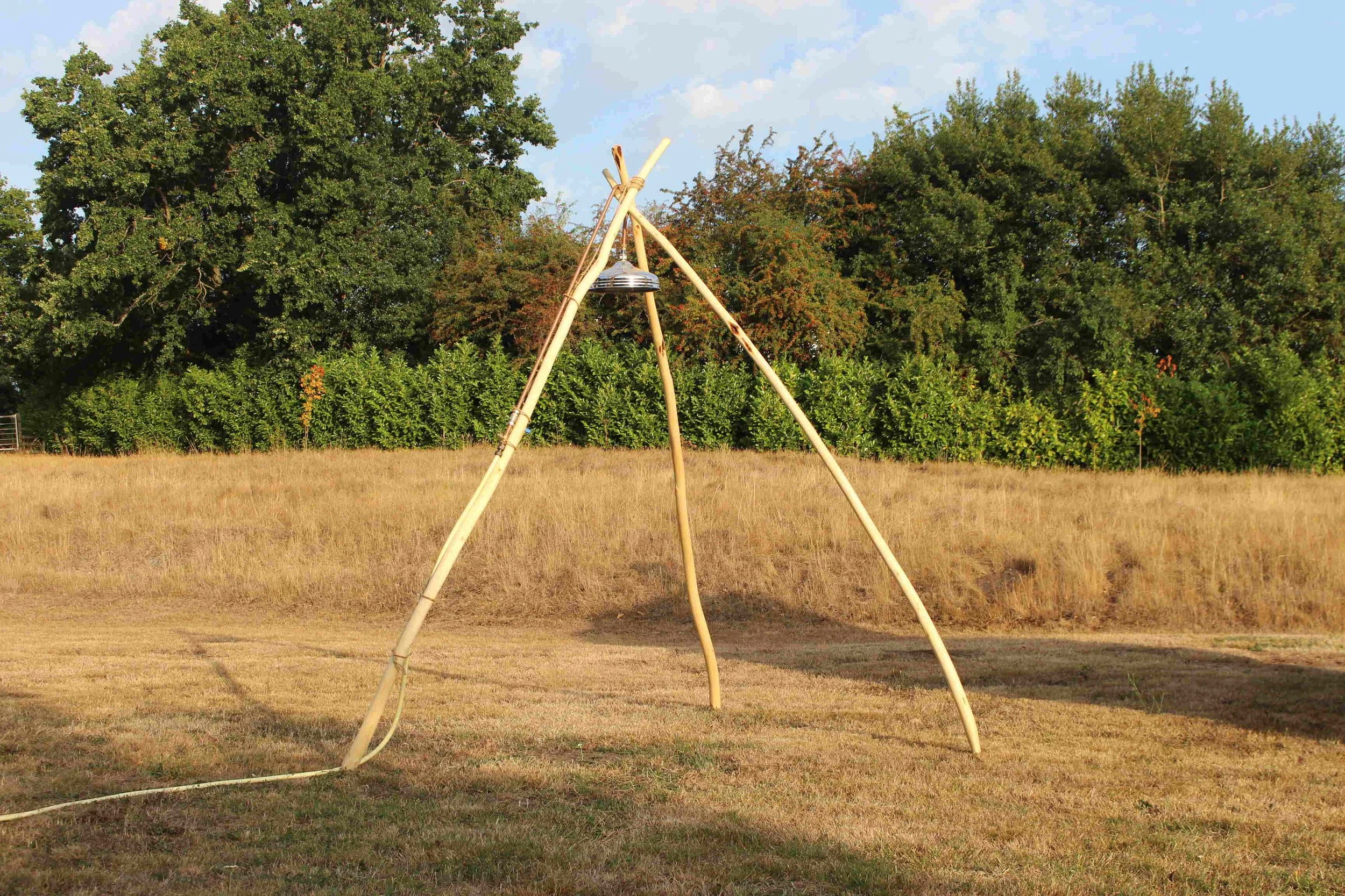 A simple wooden teepee structure with a metal bell hanging in the middle, set up on grass in an open field with trees in the background.