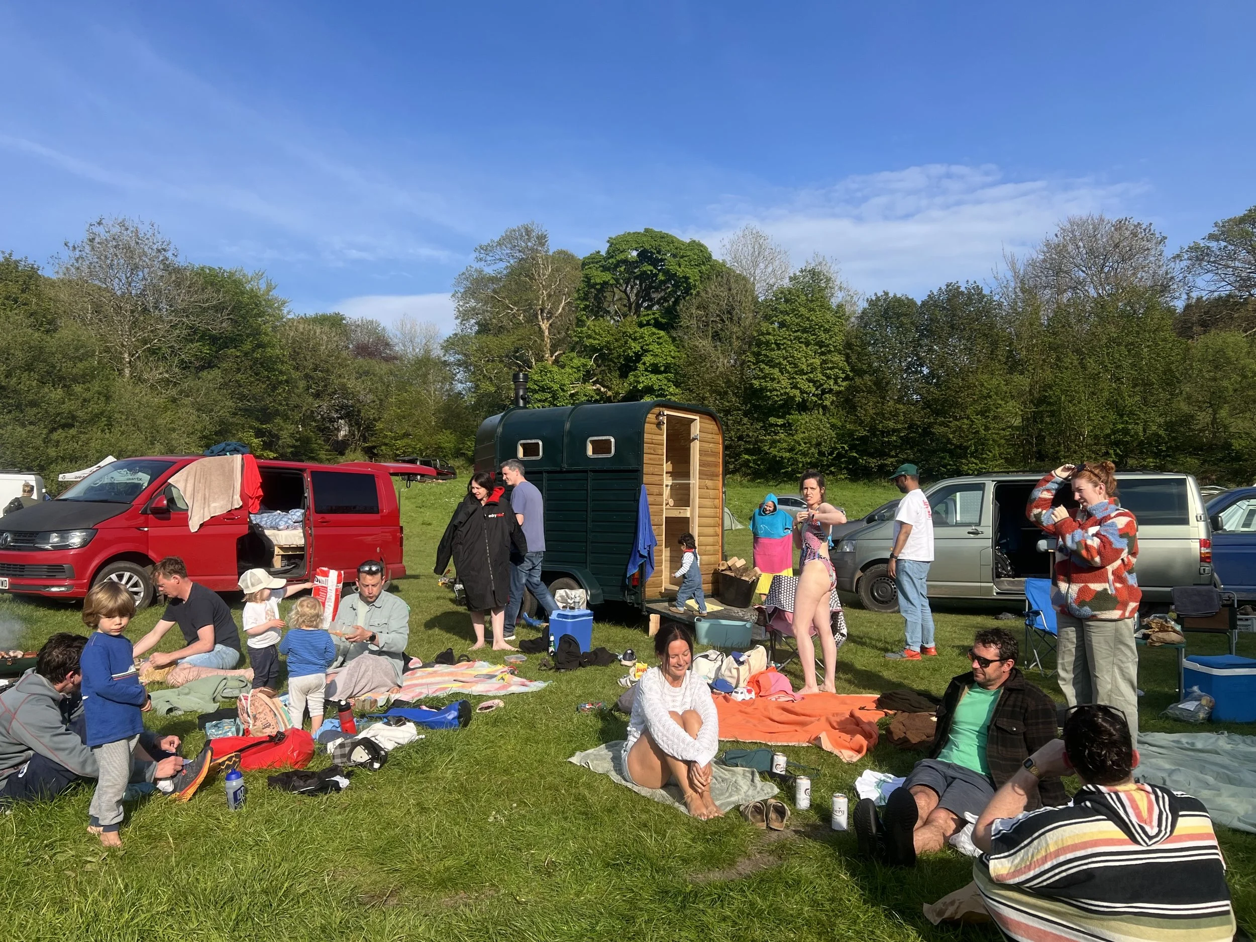 People enjoying a sunny outdoor gathering on a grassy field with trees in the background, some sitting on blankets and others standing near vans and a small wooden sauna.