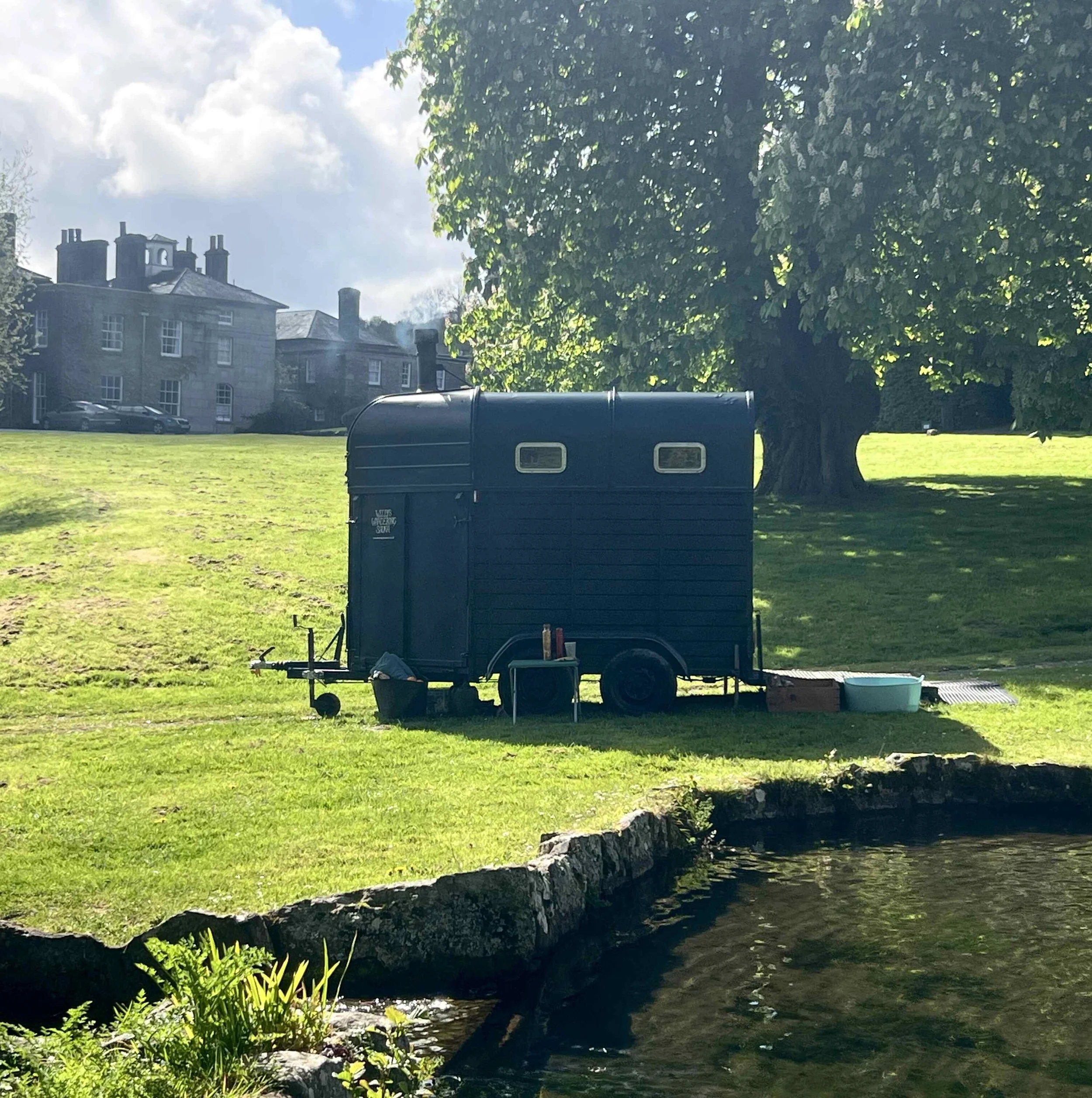A large tree with lush green leaves on the right side of the image, casting shade on the ground. In the background, there is a multi-story building with chimneys emitting smoke. In the foreground, a small body of water runs beneath a fallen tree, wit