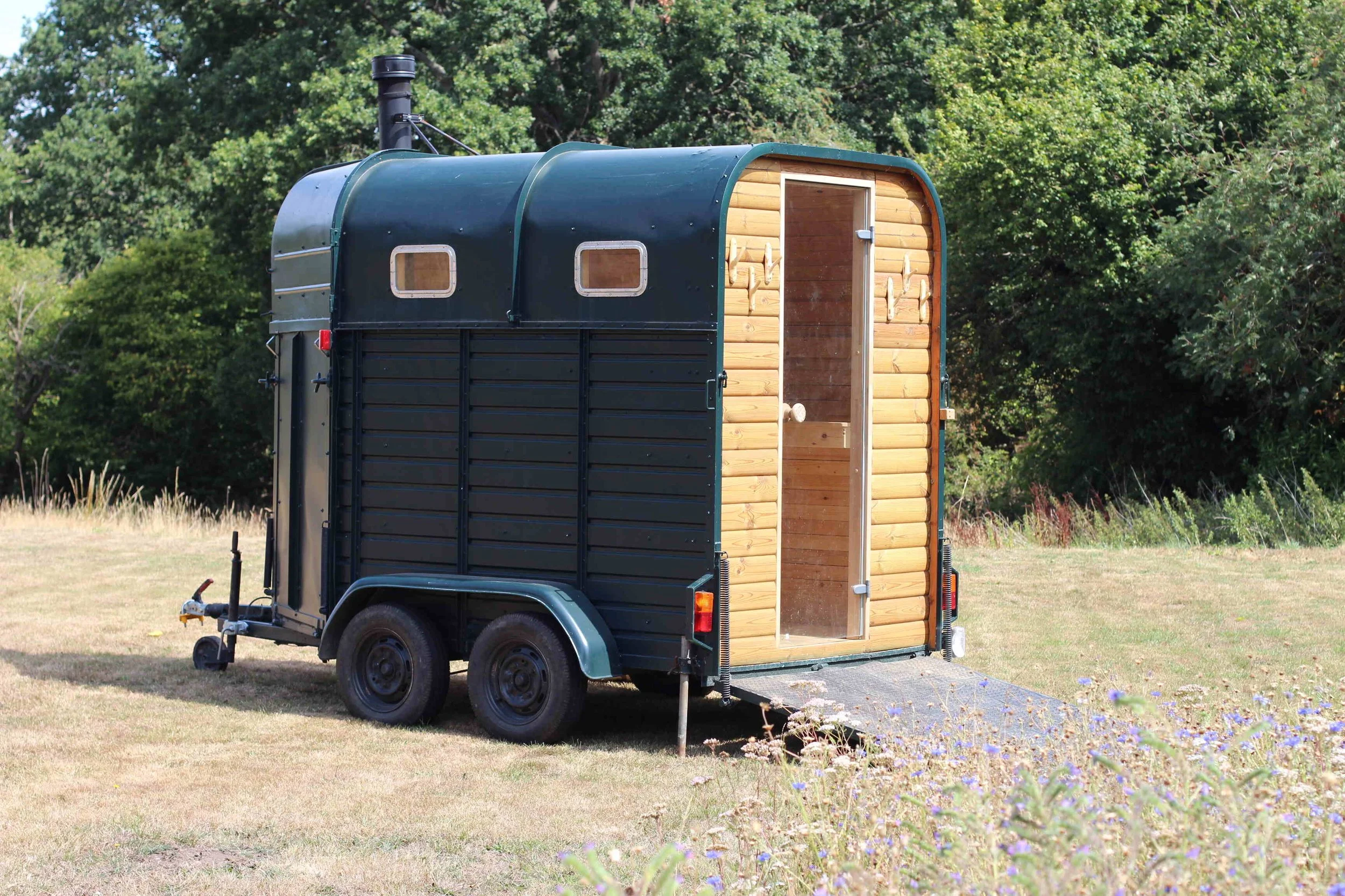 An old Rice horse box trailer converted into a mobile sauna, parked on a grassy field with trees in the background.