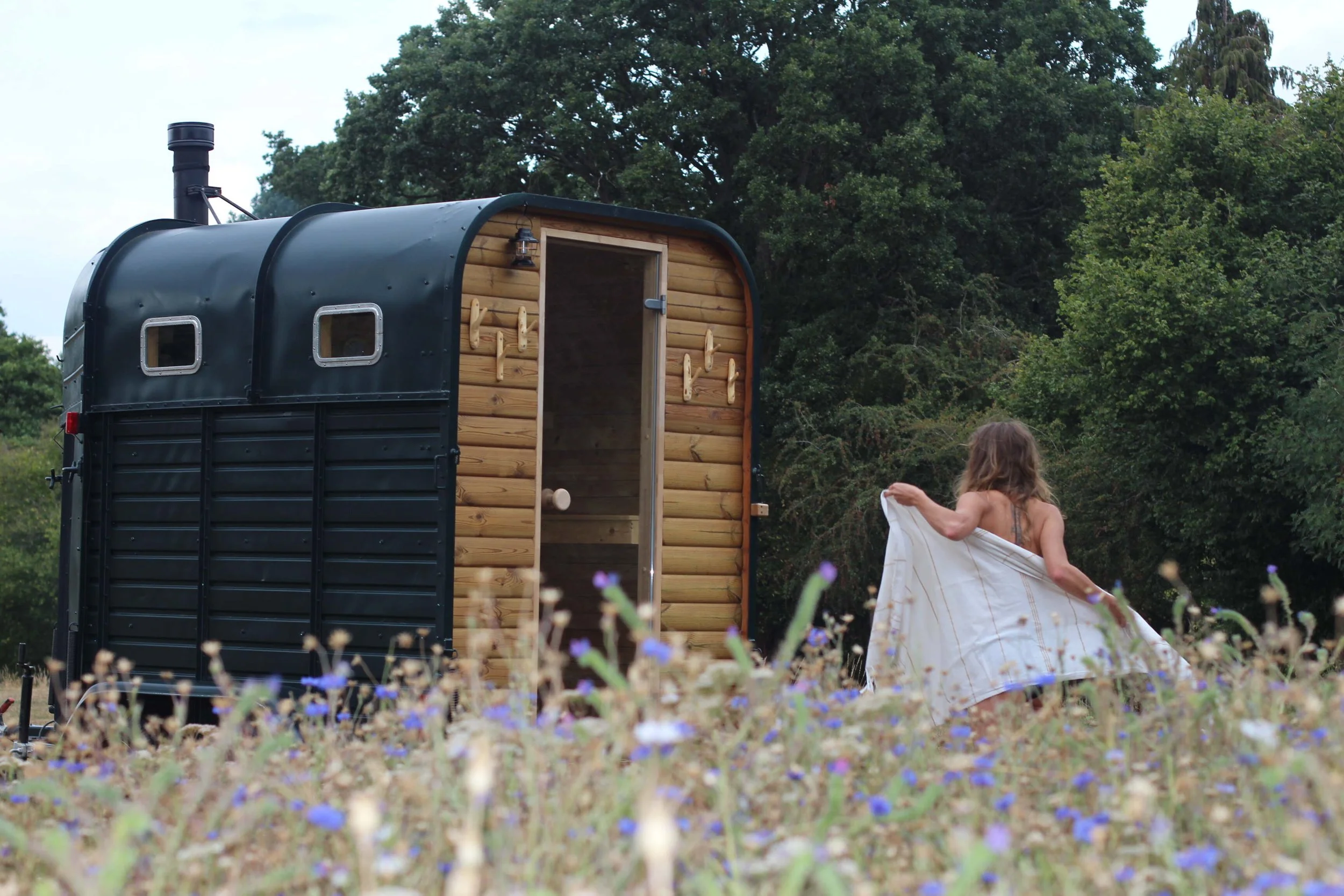 A young girl in a dress holding a white cloth in a field of wildflowers near a small black and wood-paneled camper trailer, with trees in the background.