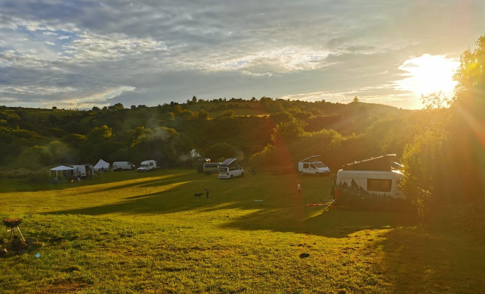 Sunset over a grassy field with several parked campervans, tents, and people, surrounded by green hills and trees. Includes Willy's Wandering Sauna, the perfect setup for a beautiful wedding in the Devon countryside.