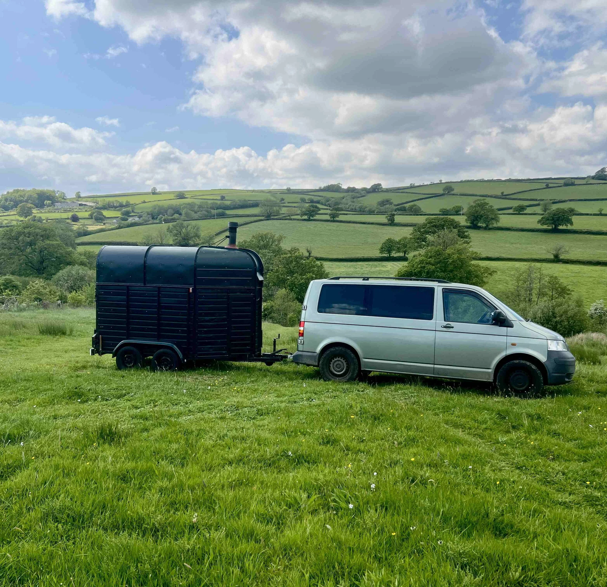 Green rolling hills with scattered trees, cloudy sky, vehicles and a trailer in the foreground.