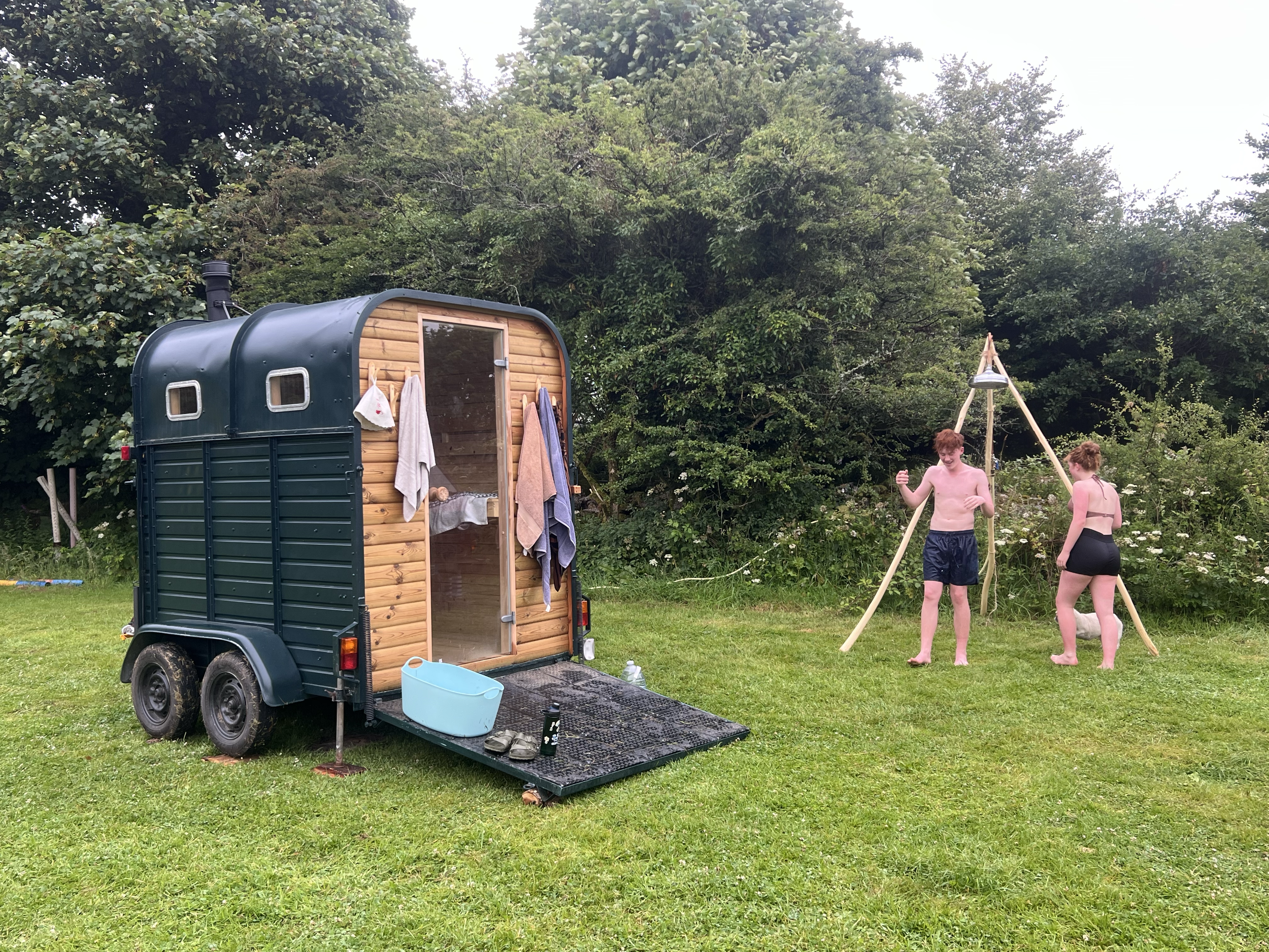 Two people in swimsuits enjoying a shower outdoors next to a tiny, dark green and wooden trailer on a grassy field with trees in the background.