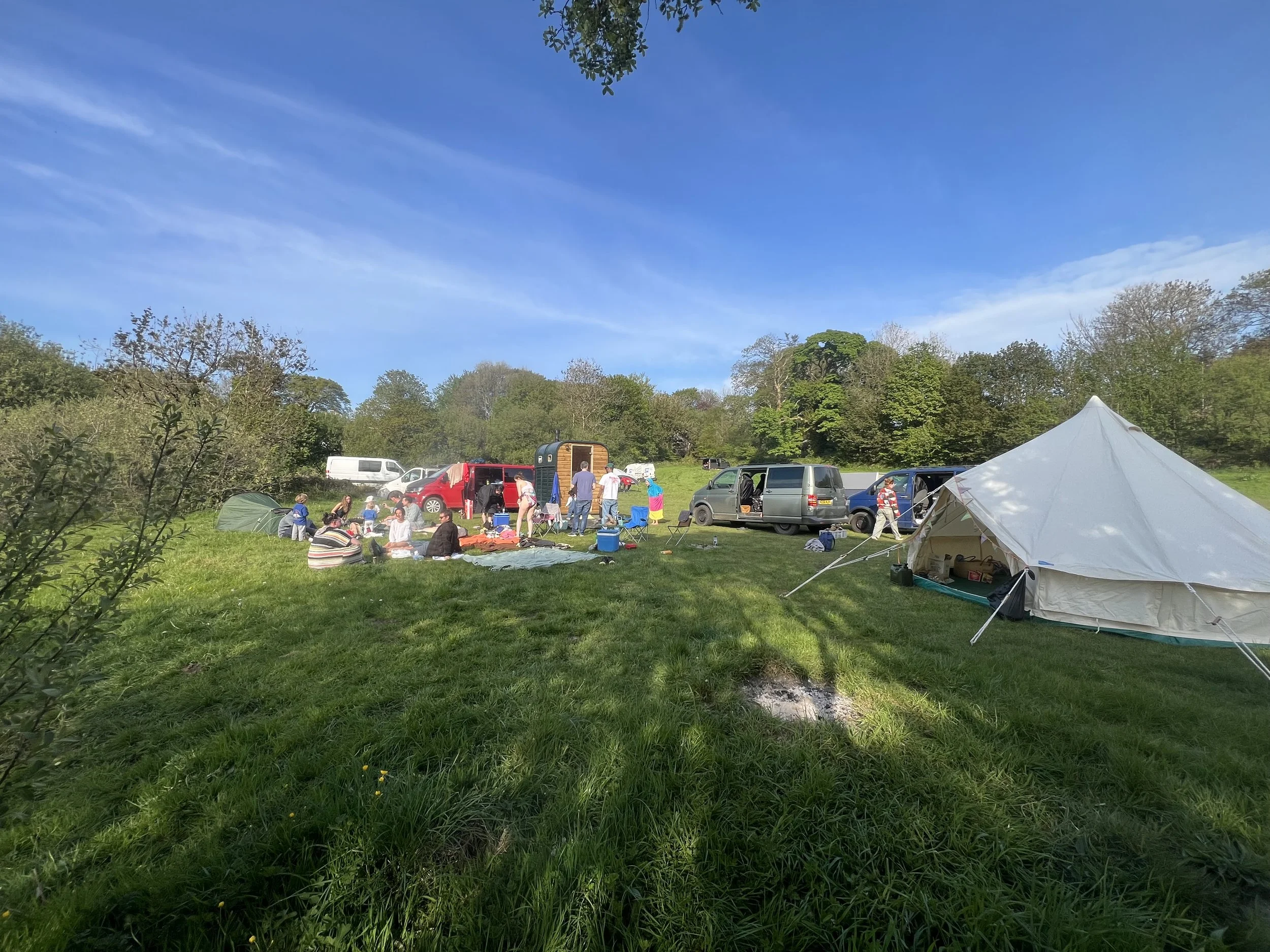 A group of people having a picnic outdoors on a grassy field in Devon under a clear blue sky with some trees in the background. There are tents, vans, and a mobile sauna set up nearby.