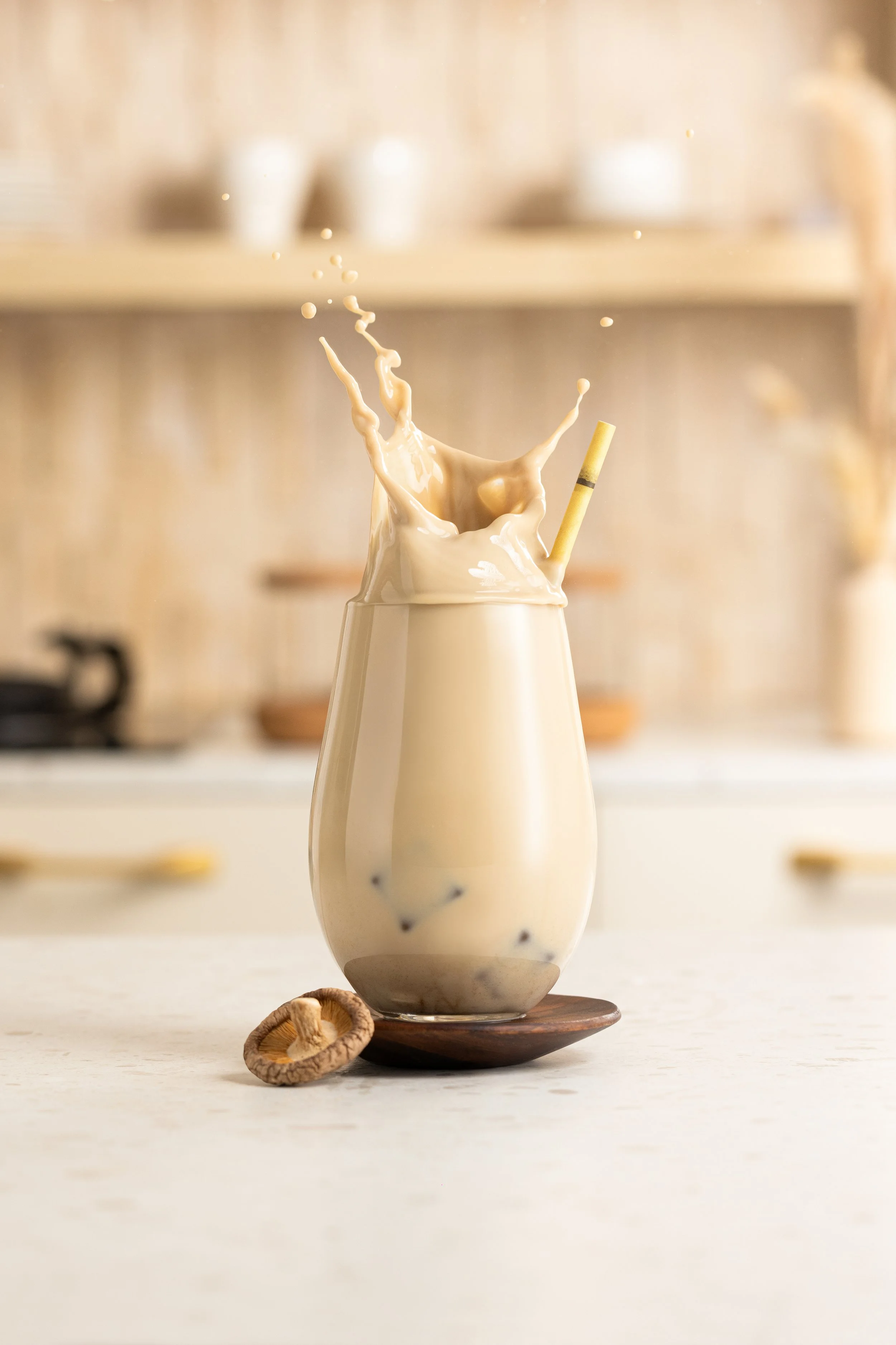A glass of bubble tea with tapioca pearls, a straw, and a splash of liquid erupting from the top, on a wooden coaster on a kitchen countertop.
