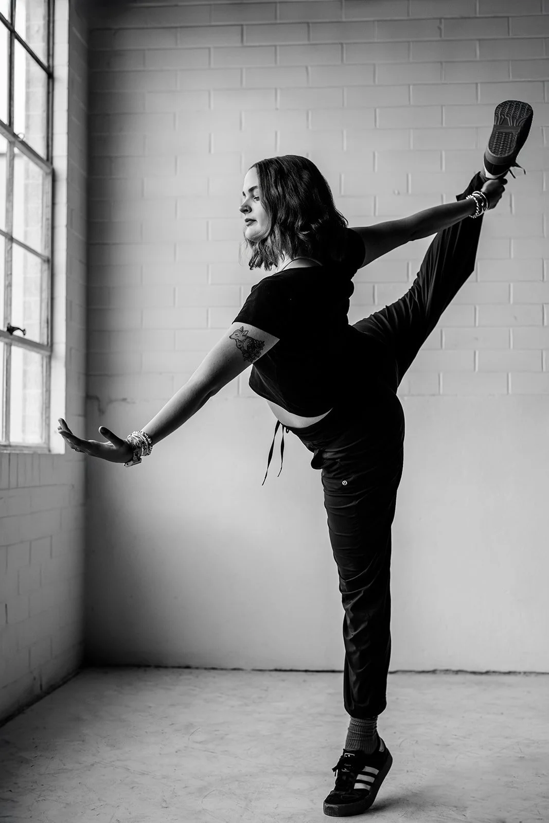 A woman with dark wavy hair practicing a high kick in a room with white brick walls and large window, wearing dark athletic clothing, sneakers, and bracelets.