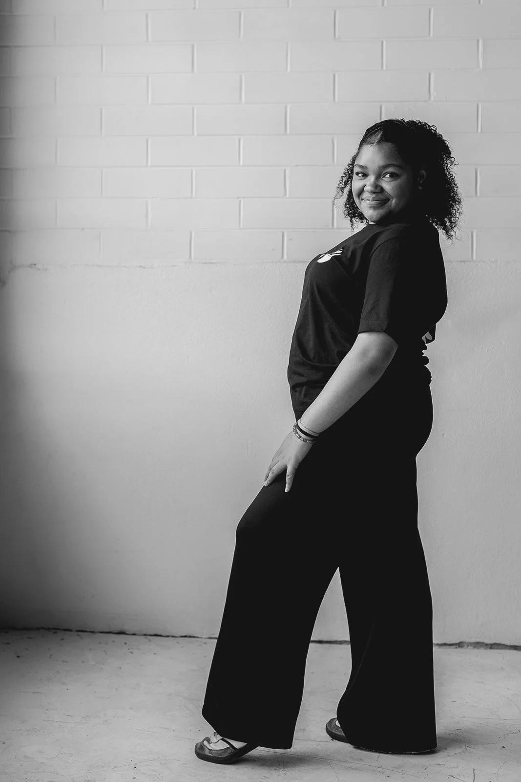 Black and white photo of a young woman smiling, standing in front of a brick wall, wearing a black t-shirt and wide-legged pants.
