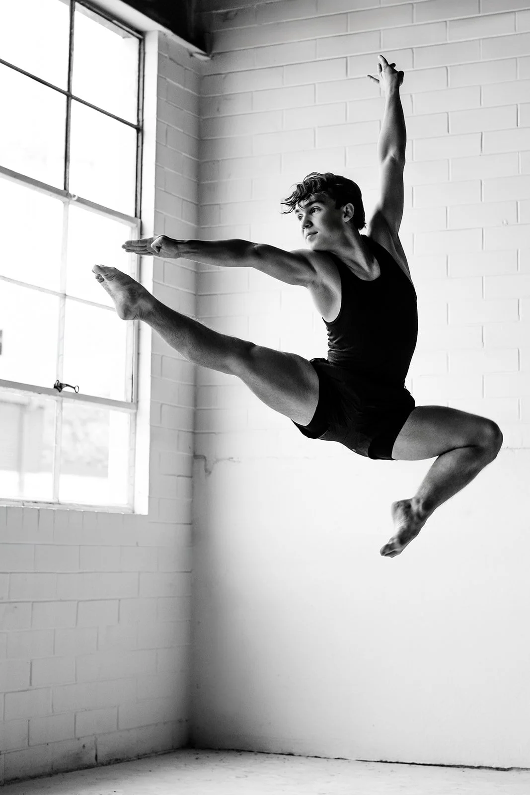 A young male dancer leaps in a studio with large windows, captured in black and white.