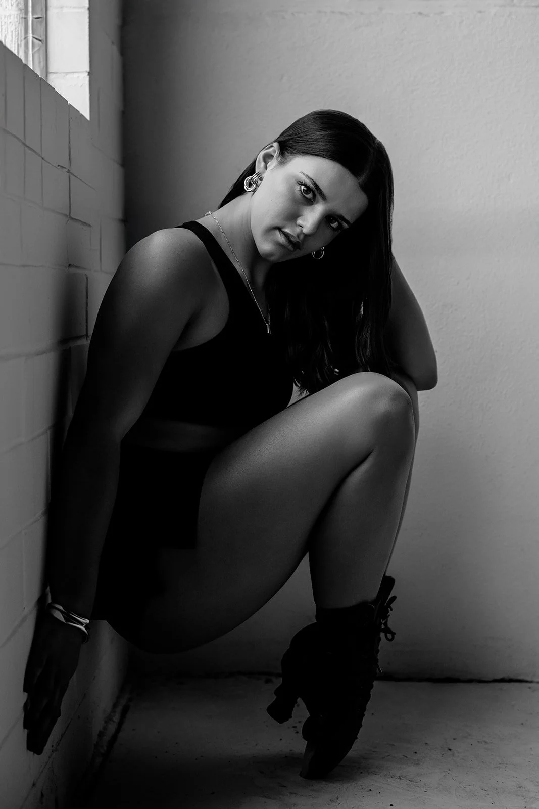 A woman with long dark hair, wearing hoop earrings, a sleeveless top, and shorts, crouches in a corner with her back against a brick wall and her knees drawn up, looking directly at the camera in black and white.